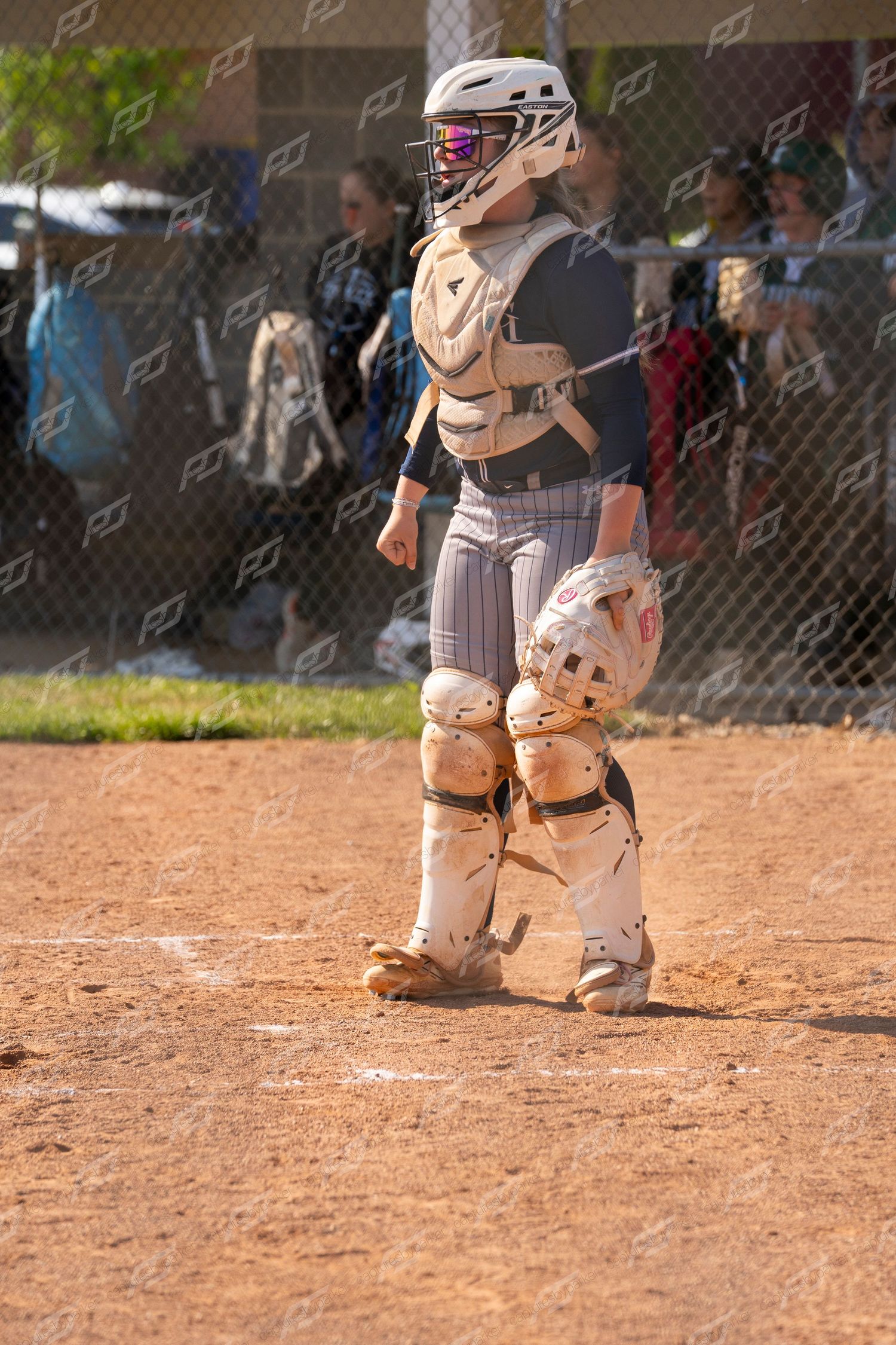 Softball catcher in full protective gear including helmet, chest protector, and shin guards stands near home plate.