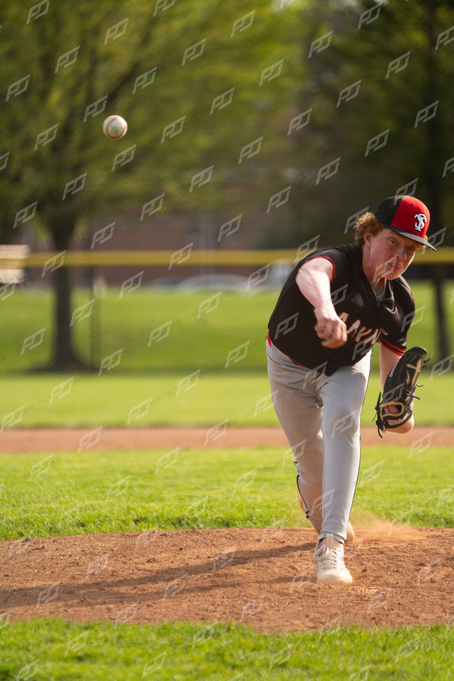 A baseball pitcher in a black uniform and red cap releases a pitch during an outdoor game on a sunny day.