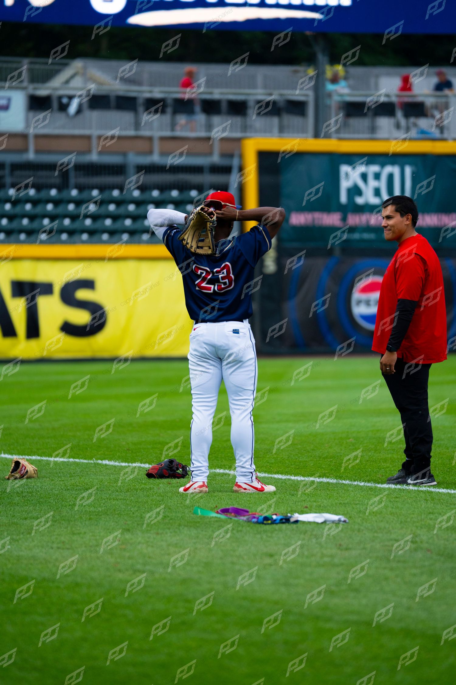 Baseball player number 23 adjusts equipment on the field while a coach watches nearby.