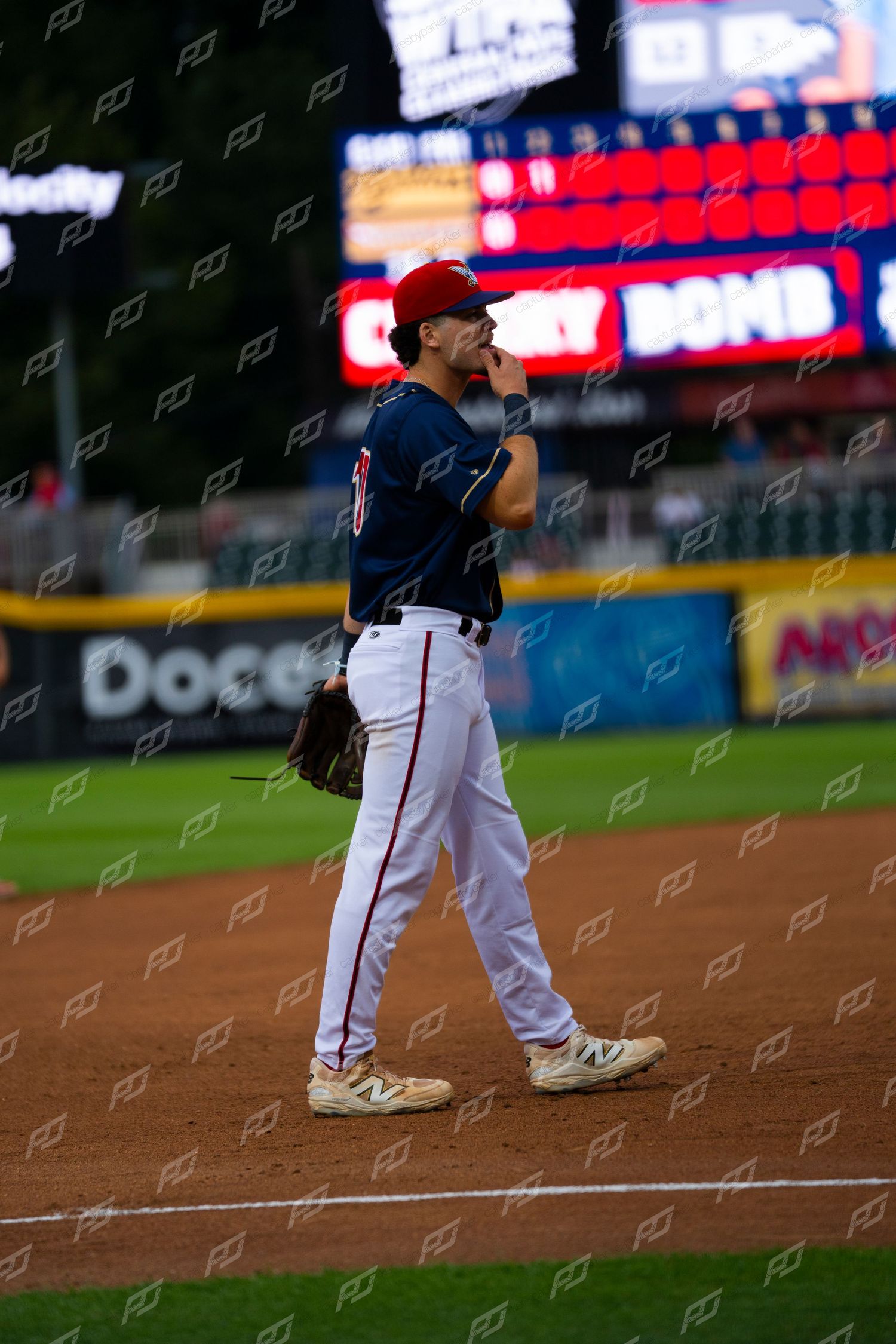 Baseball player in navy uniform stands on field with glove, scoreboard lit up behind him during night game.