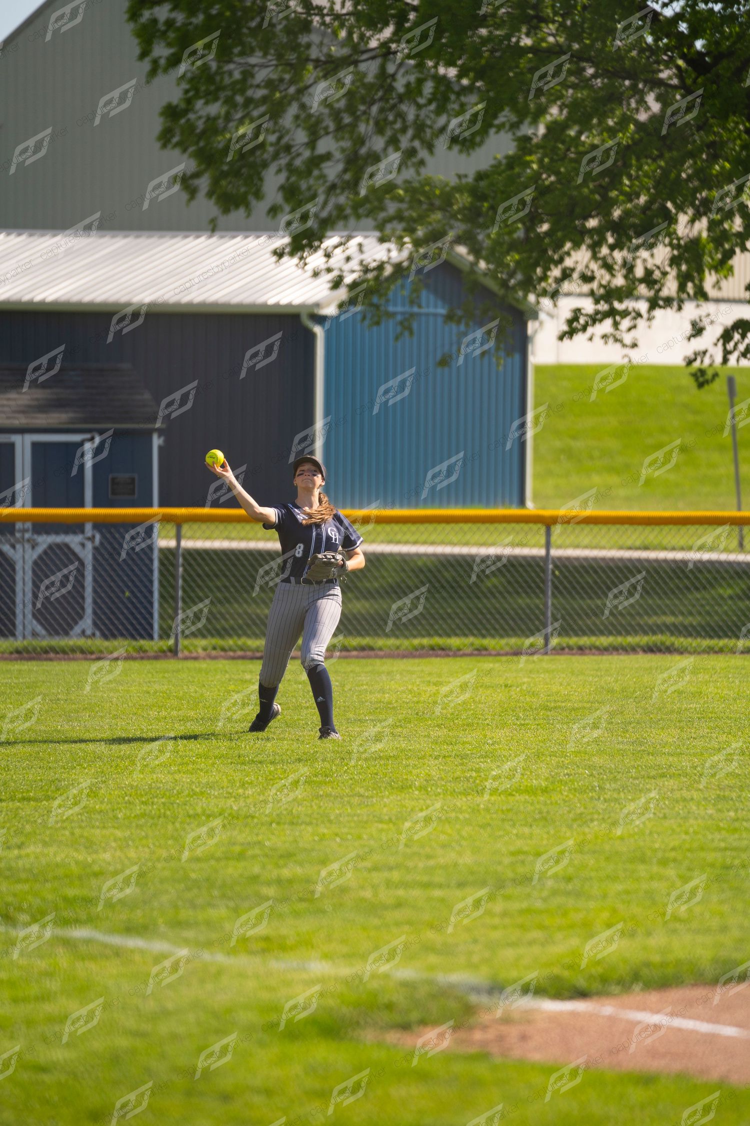 Outfielder in navy uniform reaches up to catch a softball in the outfield grass.