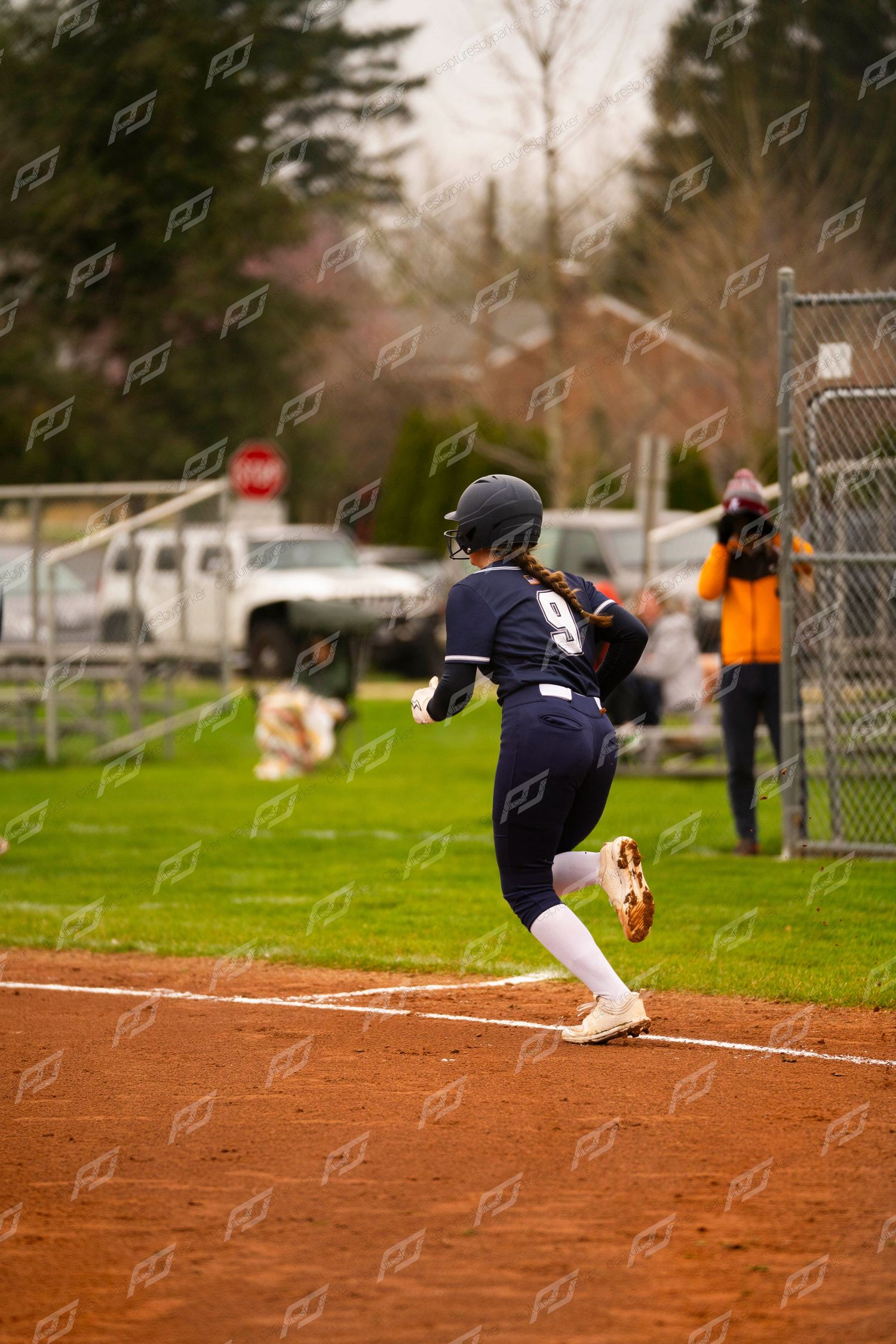 Softball player number 9 in navy uniform rounds third base while running during an outdoor game.
