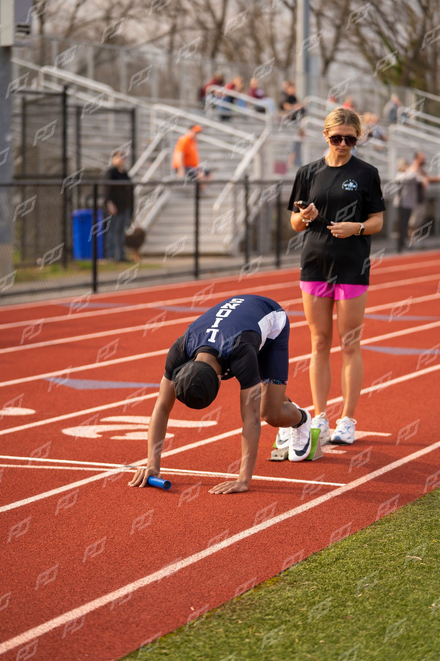 Track athlete in starting position on red track while coach observes during outdoor meet.
