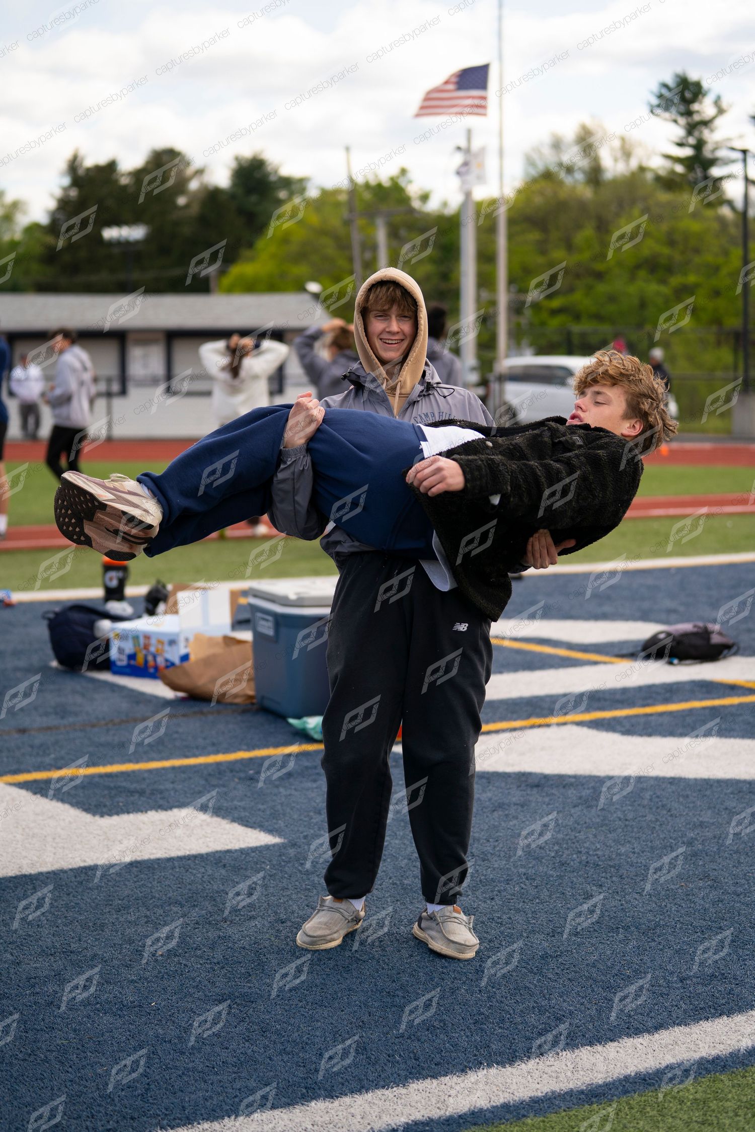 A smiling athlete carries a teammate in his arms on a blue turf football field at a track and field meet.