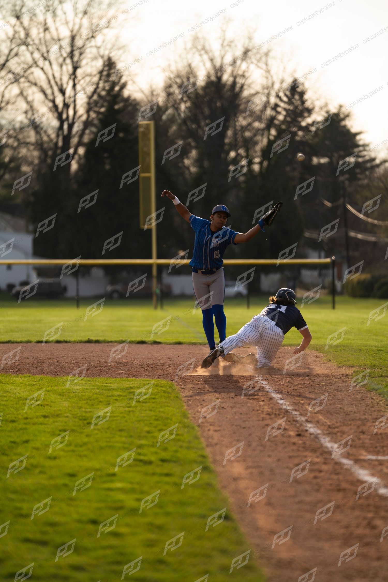 Baseball player slides into base as opposing fielder in blue uniform leaps to catch the ball.