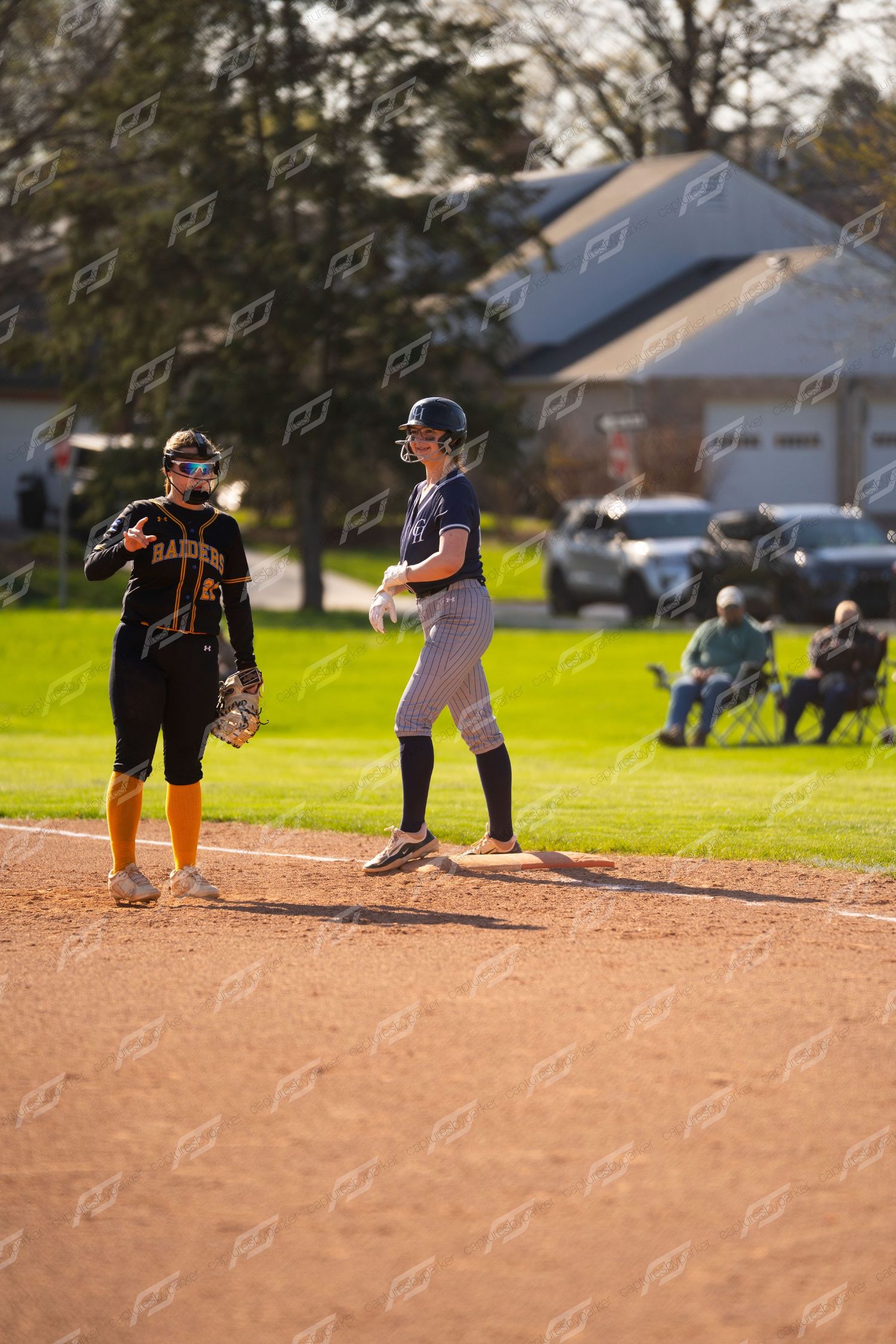 Female softball player in navy uniform stands on base next to opponent wearing Raiders jersey.