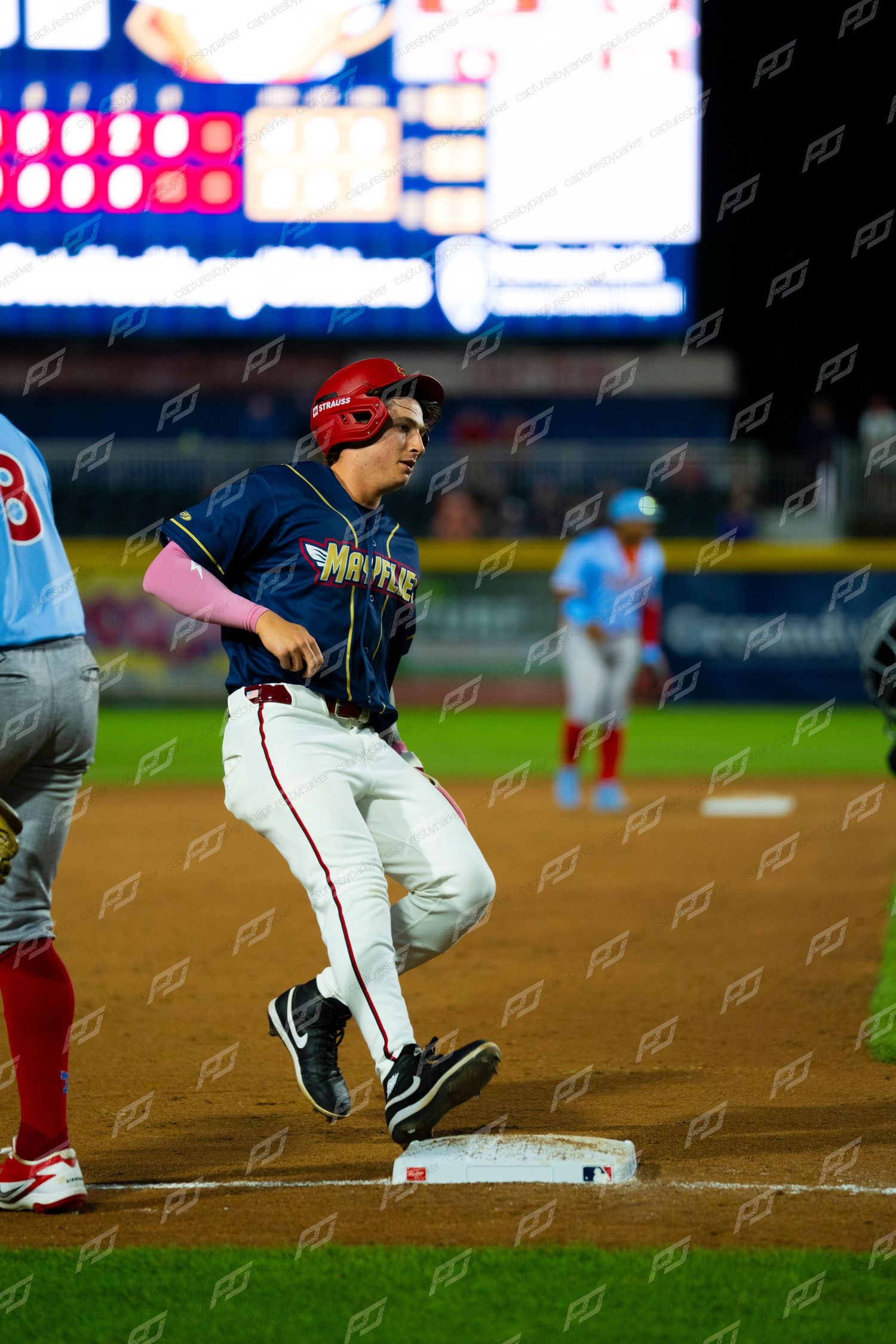 Baseball player rounds third base during a night game with scoreboard and teammates visible.