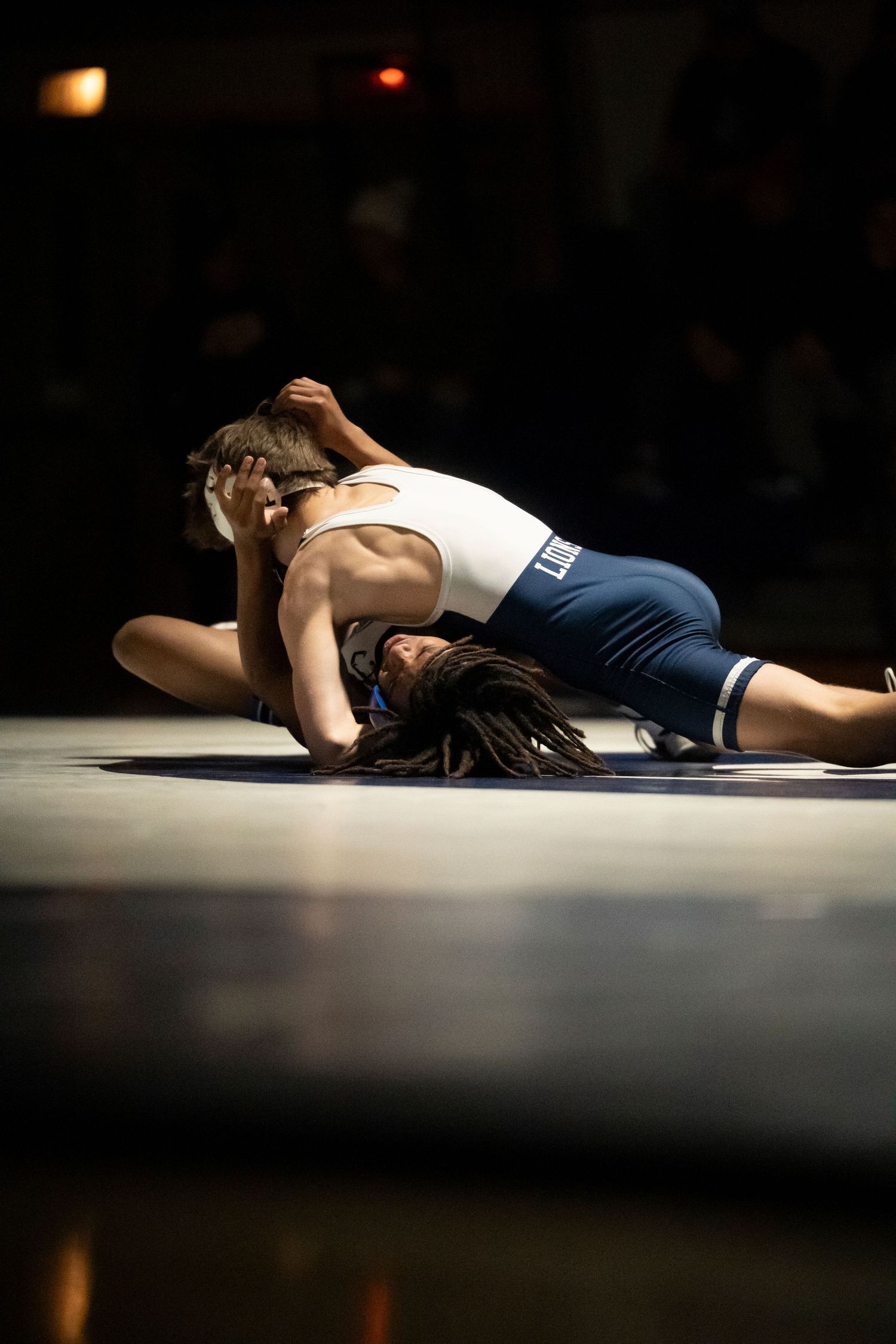 A wrestler in white pins his dreadlocked opponent to the mat in a dramatic wrestling match under dark lighting.