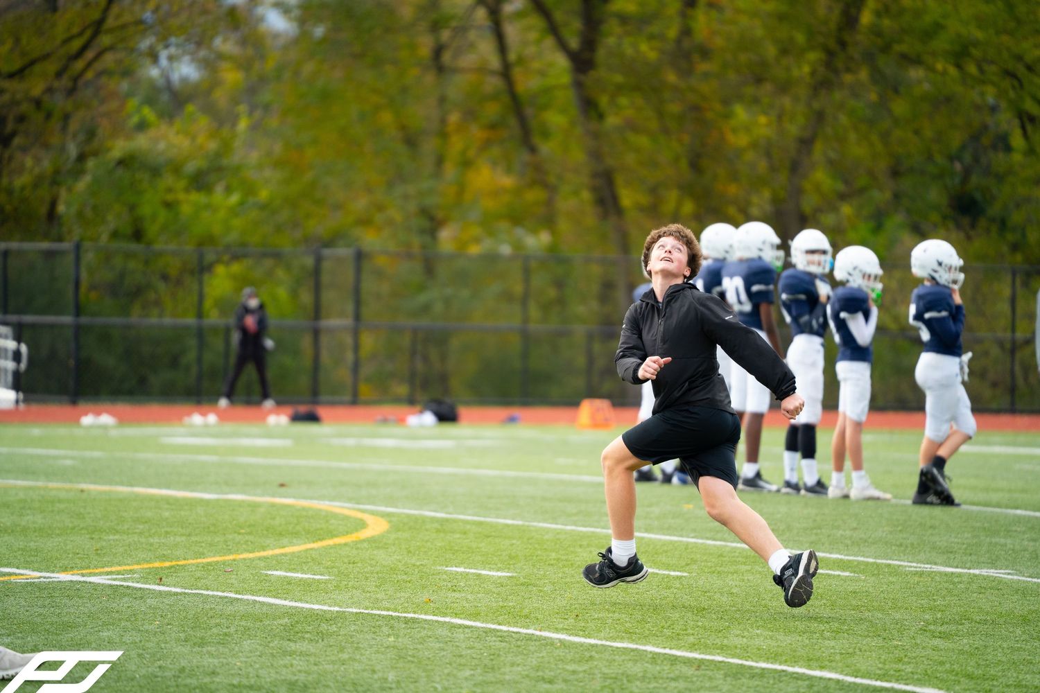 Coach in black jacket runs energetically on green turf football field.