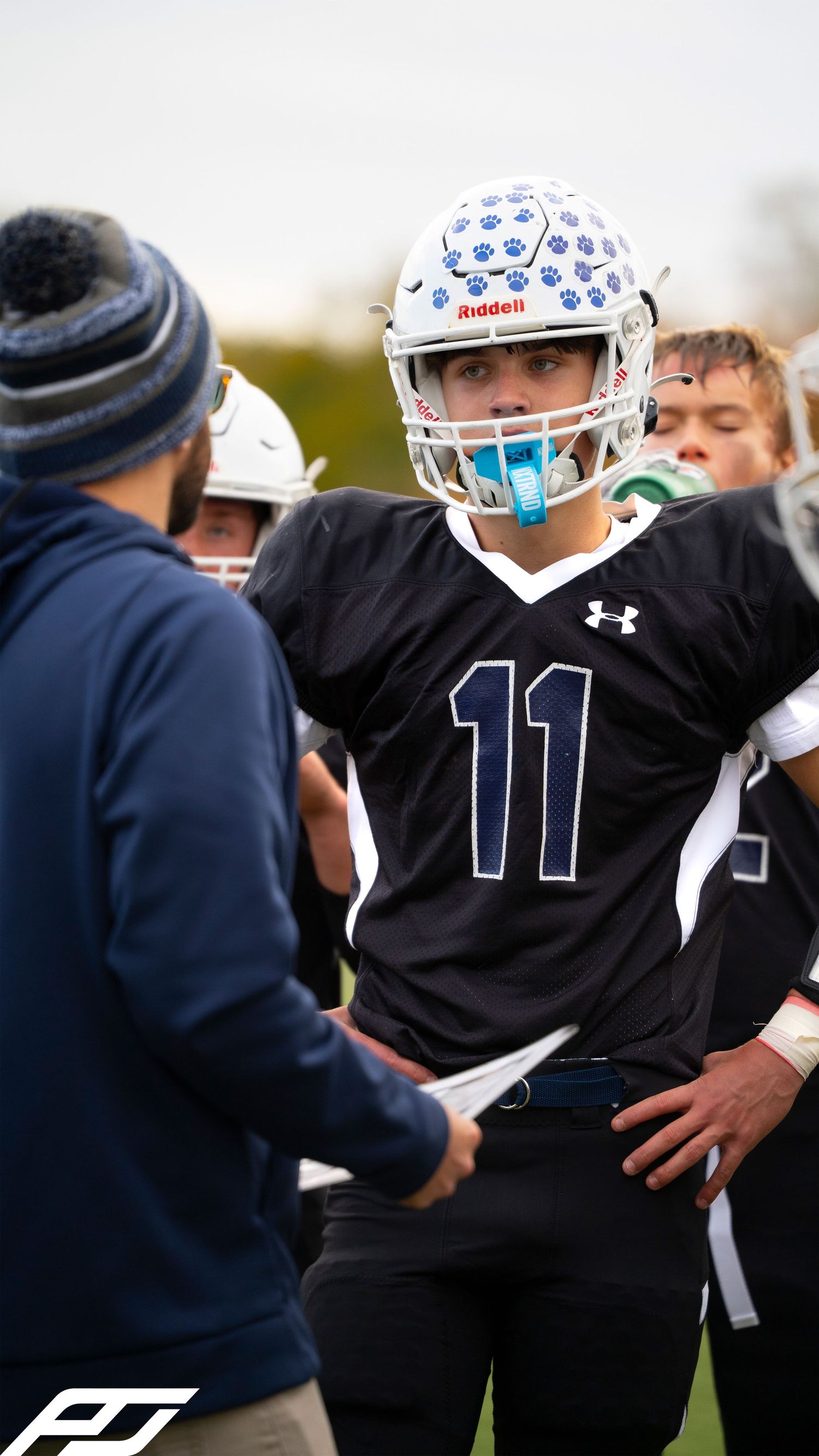 Youth football player #11 in black Under Armour uniform listens to coach during game.