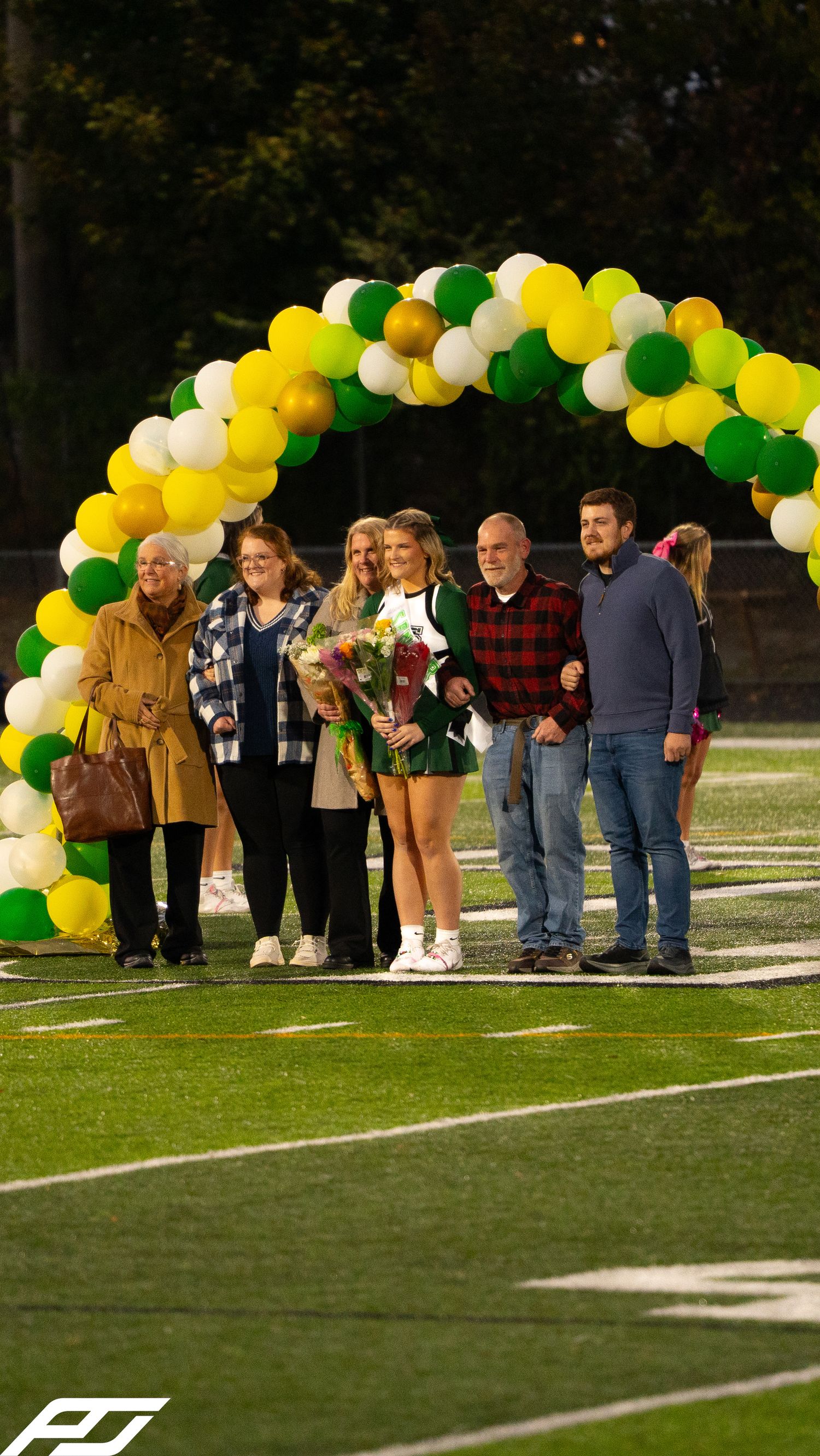 Cheerleader senior night celebration with large family group under green and gold balloon arch on football field.