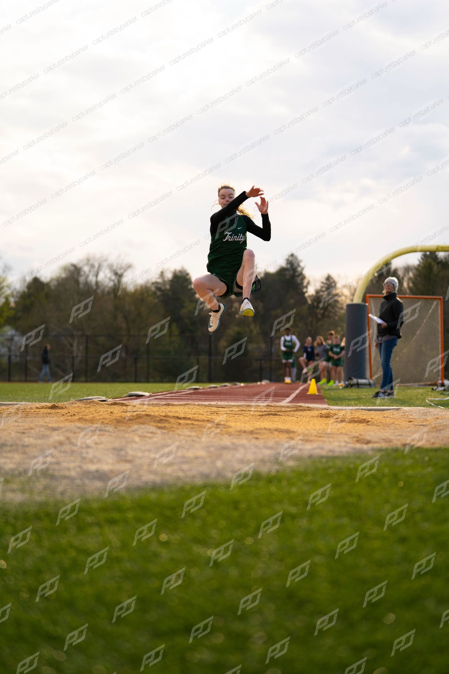 High school athlete in green uniform soars through air during long jump competition at track meet.