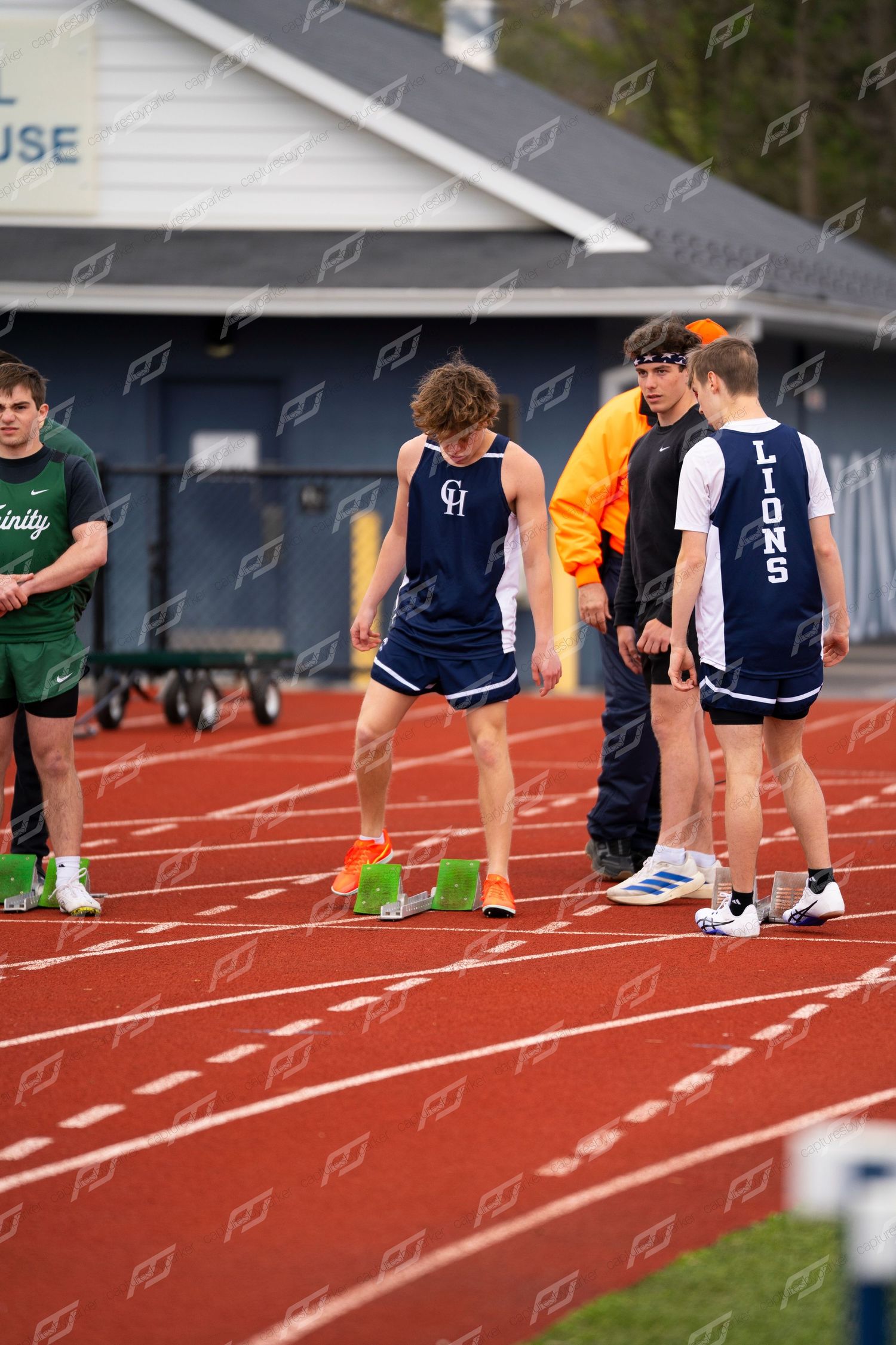 High school male track athletes in GH and Lions uniforms prepare at starting line on red track.