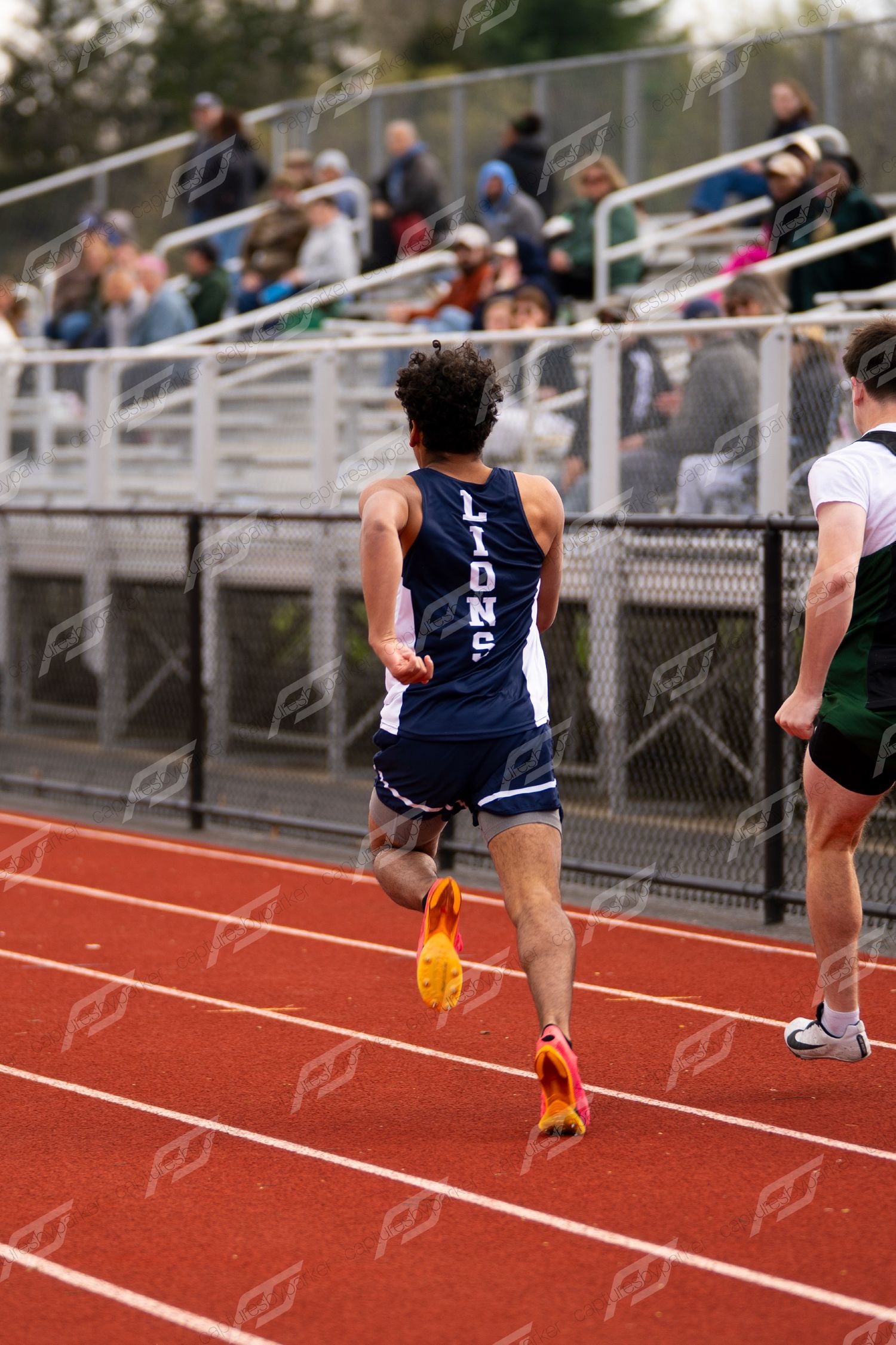 Lions track athlete runs from behind on red track with spectators in bleachers at high school meet.