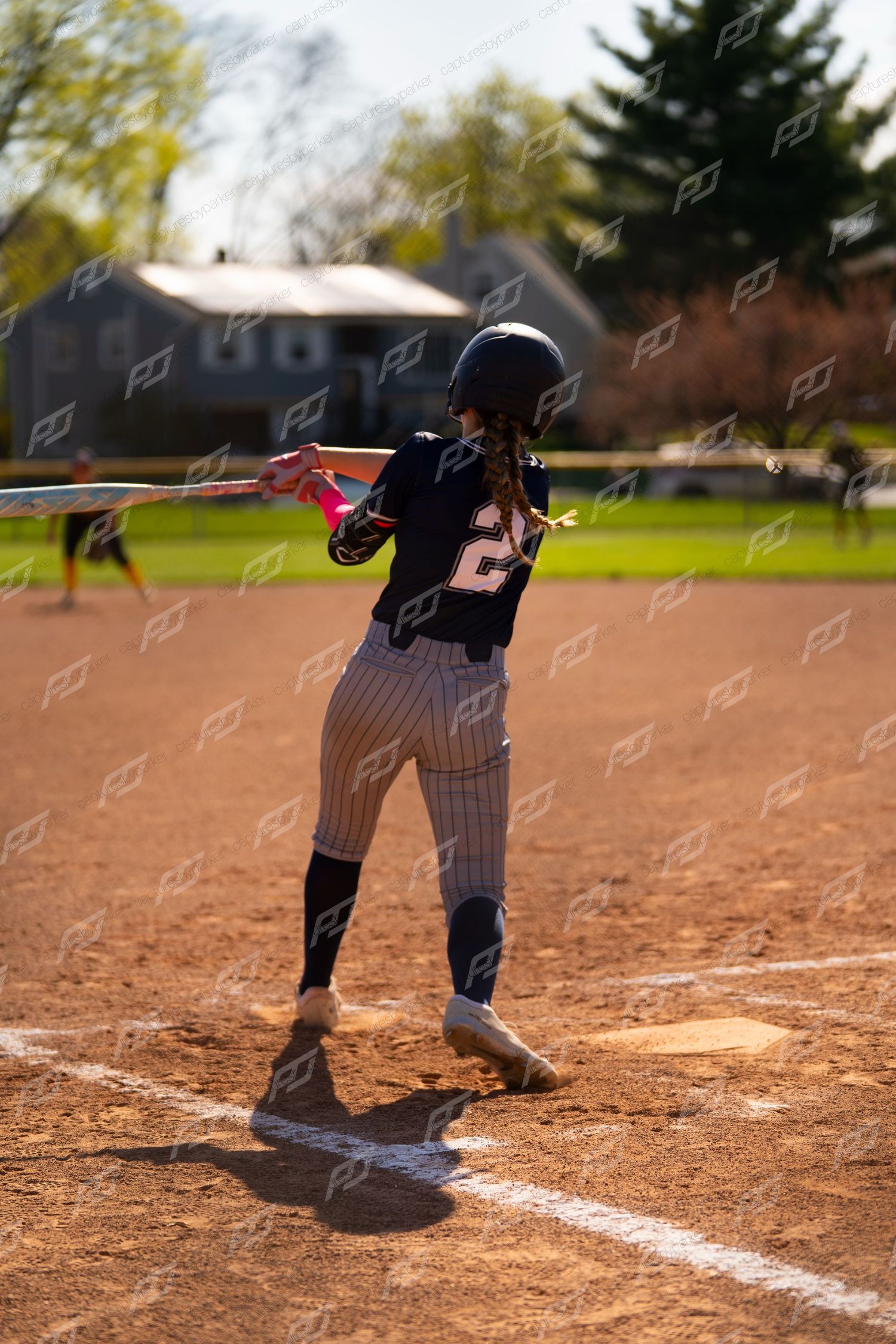 Softball player number 2 in navy pinstripe uniform swings bat at home plate during outdoor game.