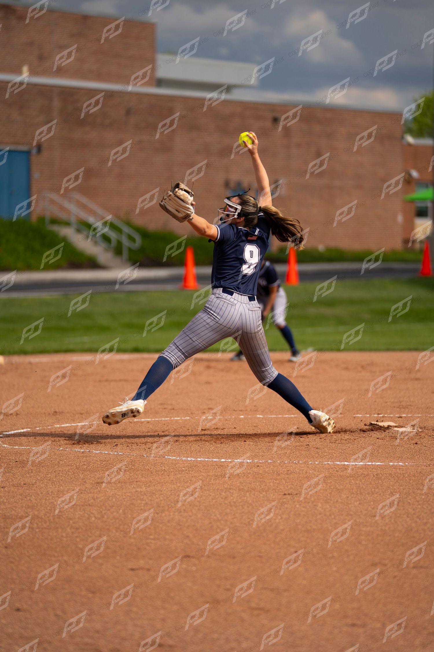 Female softball pitcher in navy uniform winding up to deliver an underhand pitch.