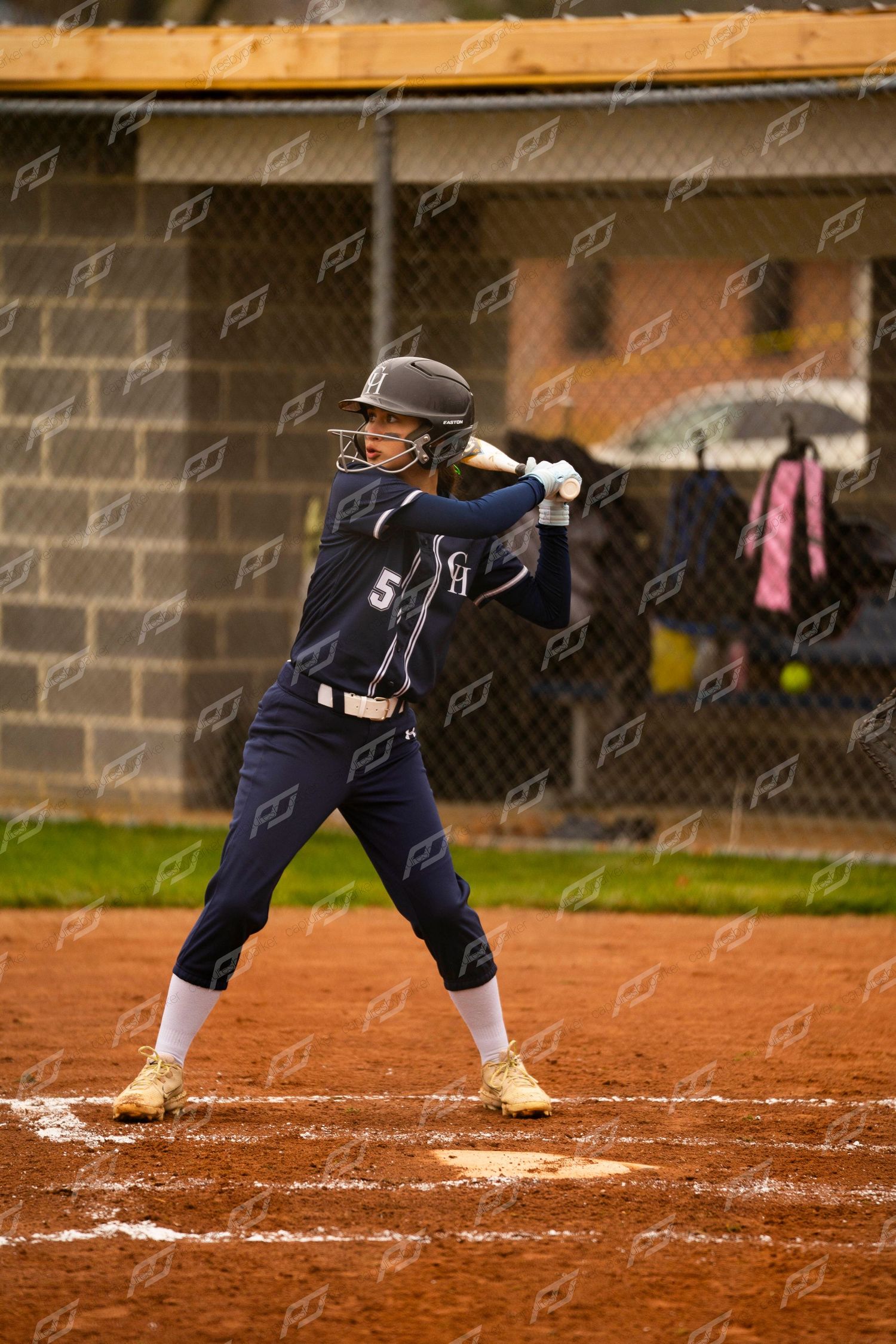 Female softball player in navy #5 jersey stands ready in batting stance at home plate.