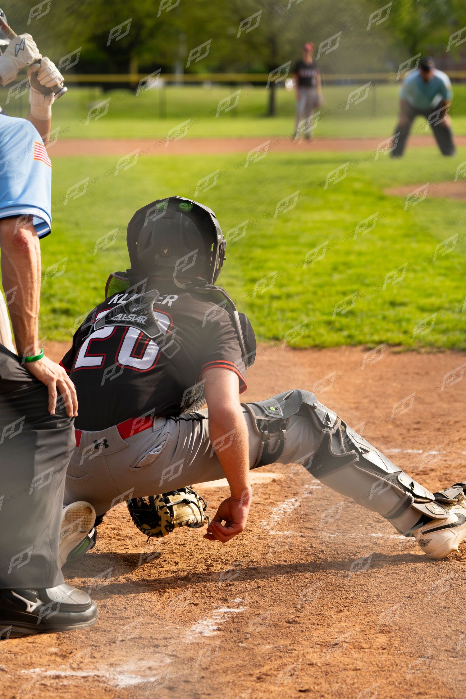 Baseball catcher number 20 in black uniform crouches low to field a pitch during a sunny game.