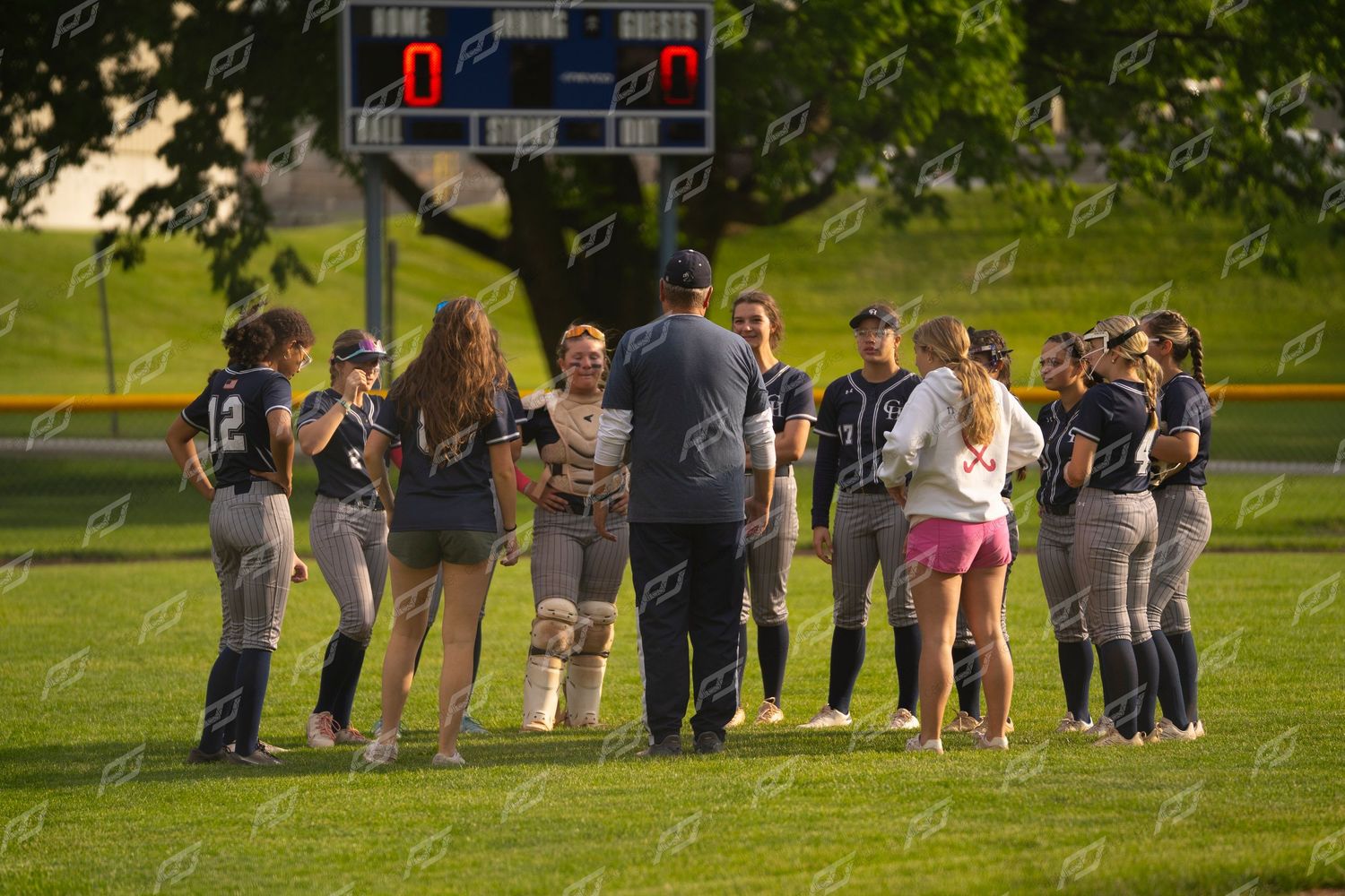 Softball team in navy uniforms huddles with coach near scoreboard after game.