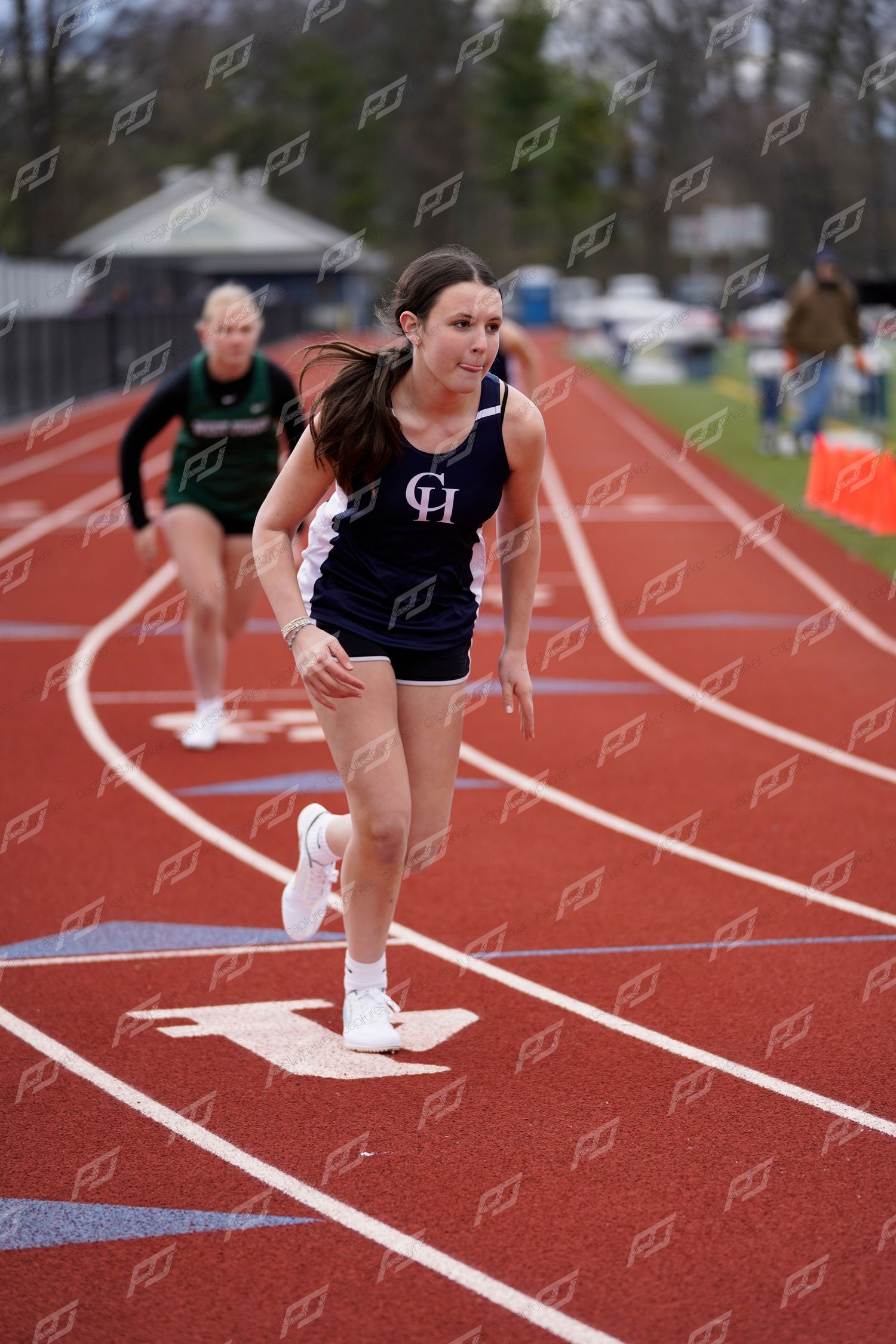 Female track athlete in navy CH uniform sprints forward on red track during a competitive meet.