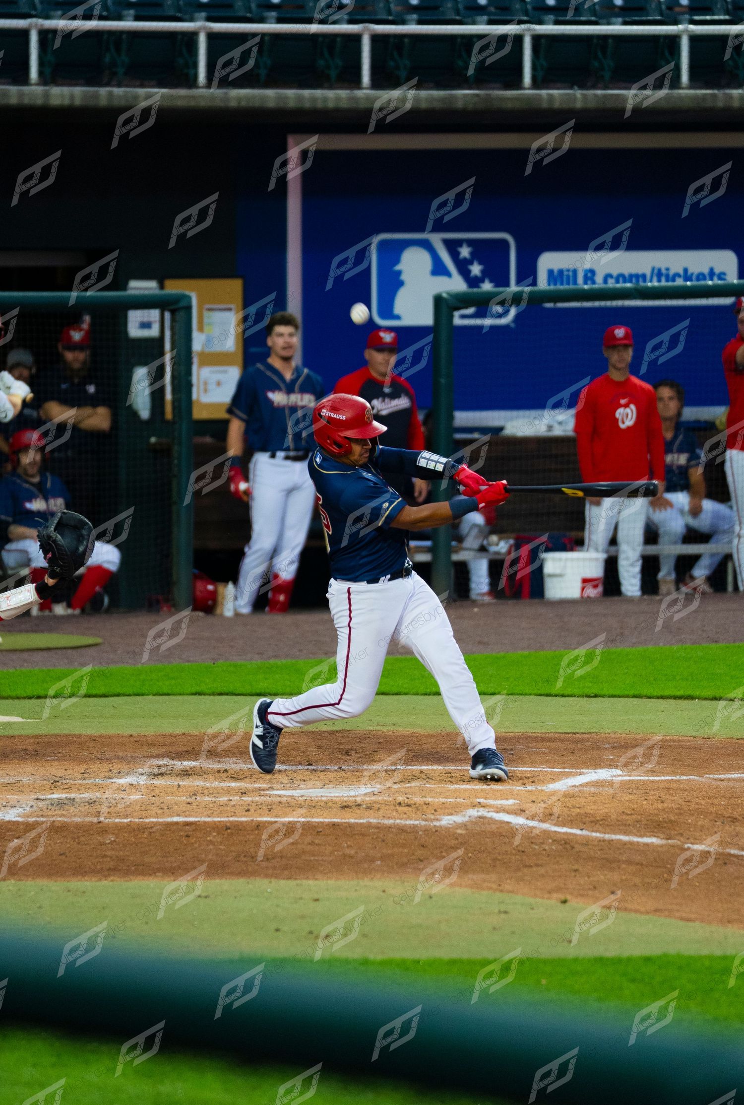 Minor league batter in red helmet swings at pitch near dugout during evening baseball game.