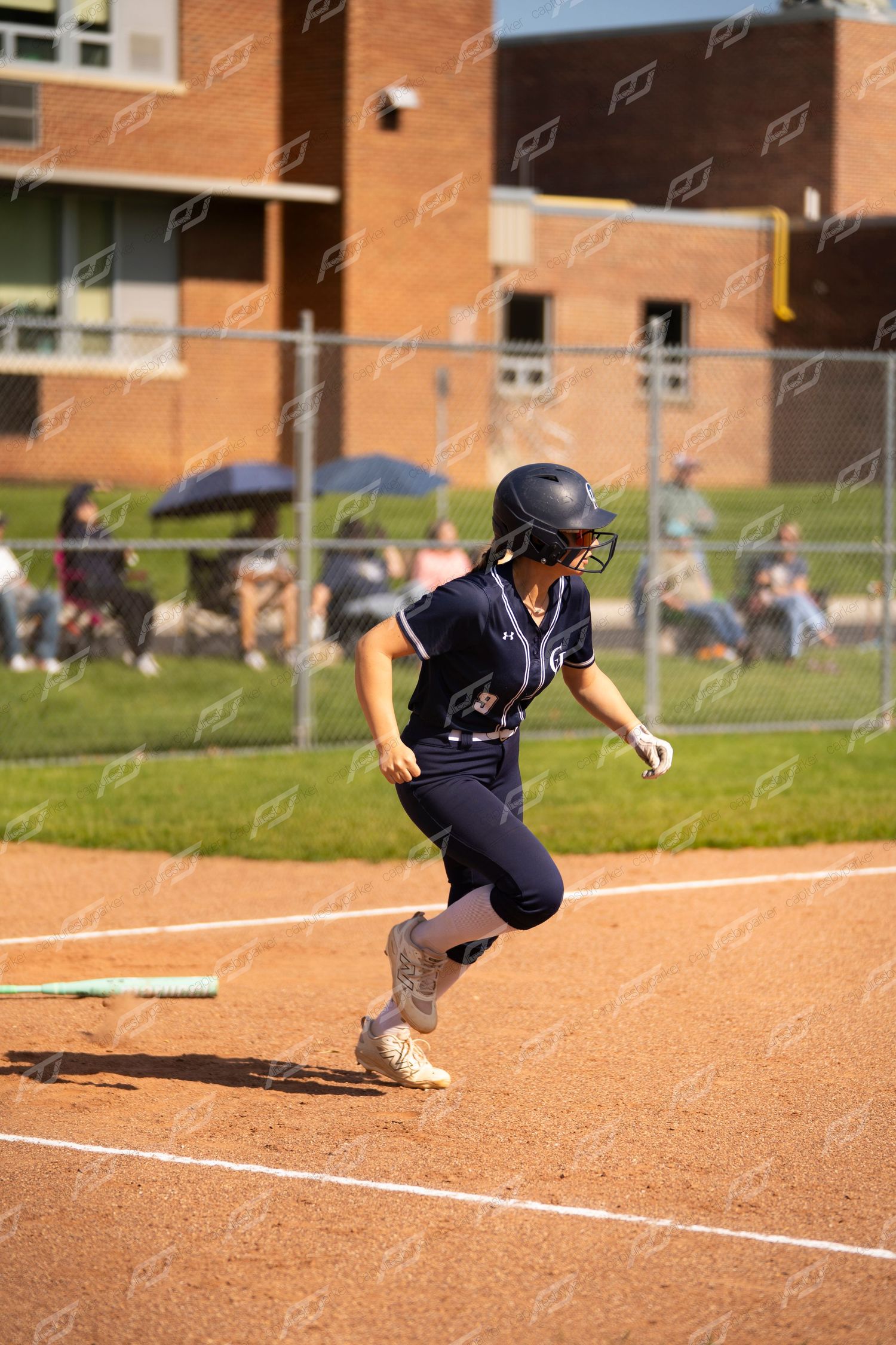 Female softball player in navy uniform sprints along baseline after making contact at home plate.
