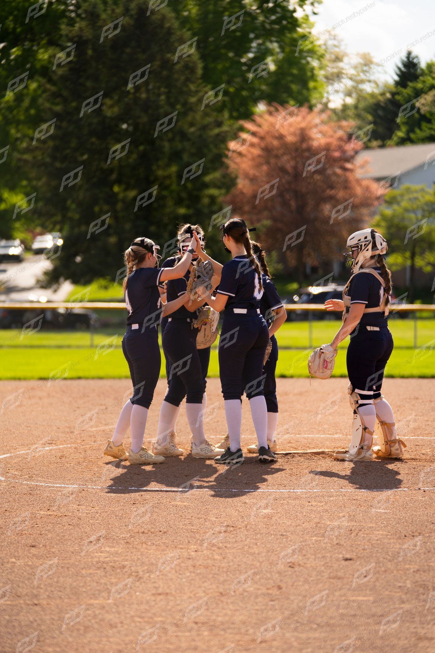 Four softball players in navy uniforms huddle near the pitcher's mound during a game.