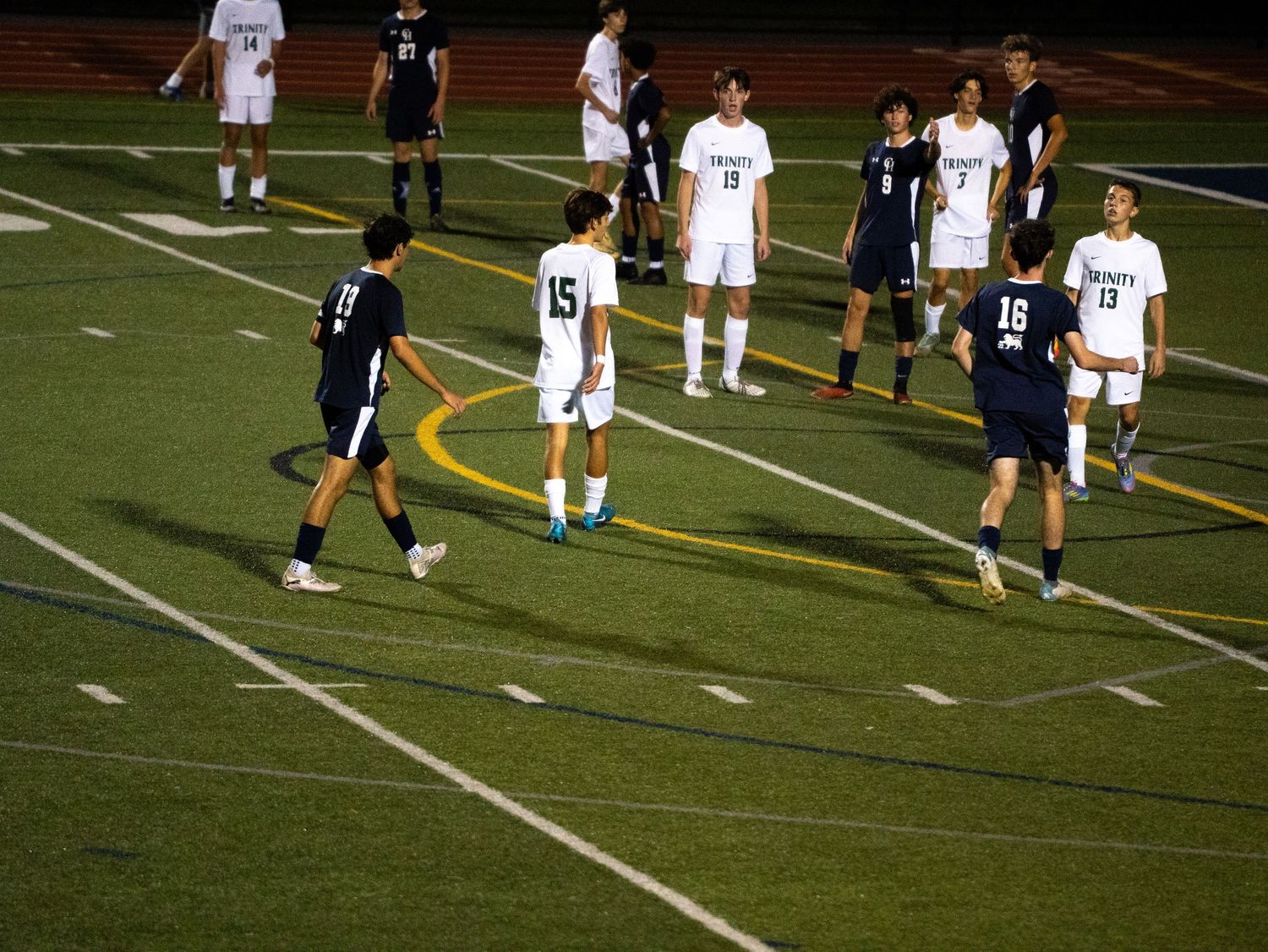High school soccer players in navy and white uniforms position on lit turf field at night.