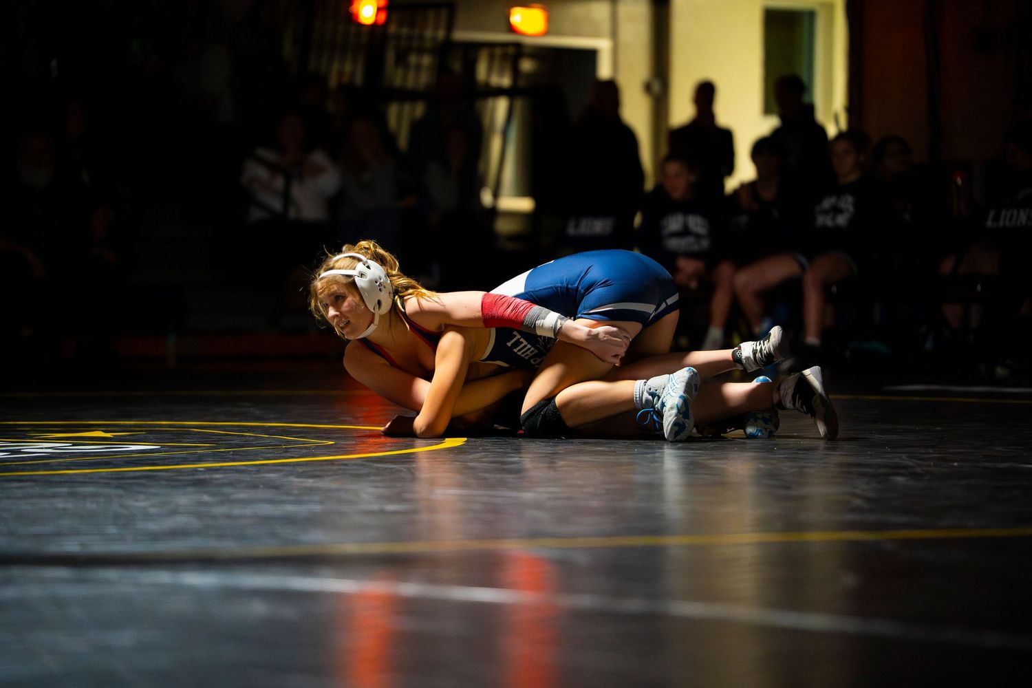 Female wrestler in blue uniform pins opponent to yellow wrestling mat during competition.