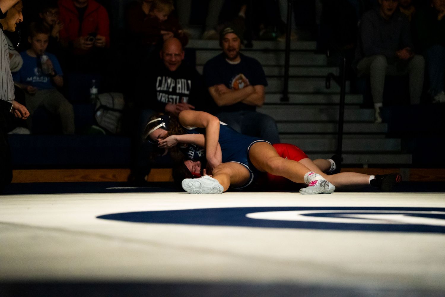 Wrestler in blue singlet executes takedown move on opponent in red during competitive high school wrestling match.