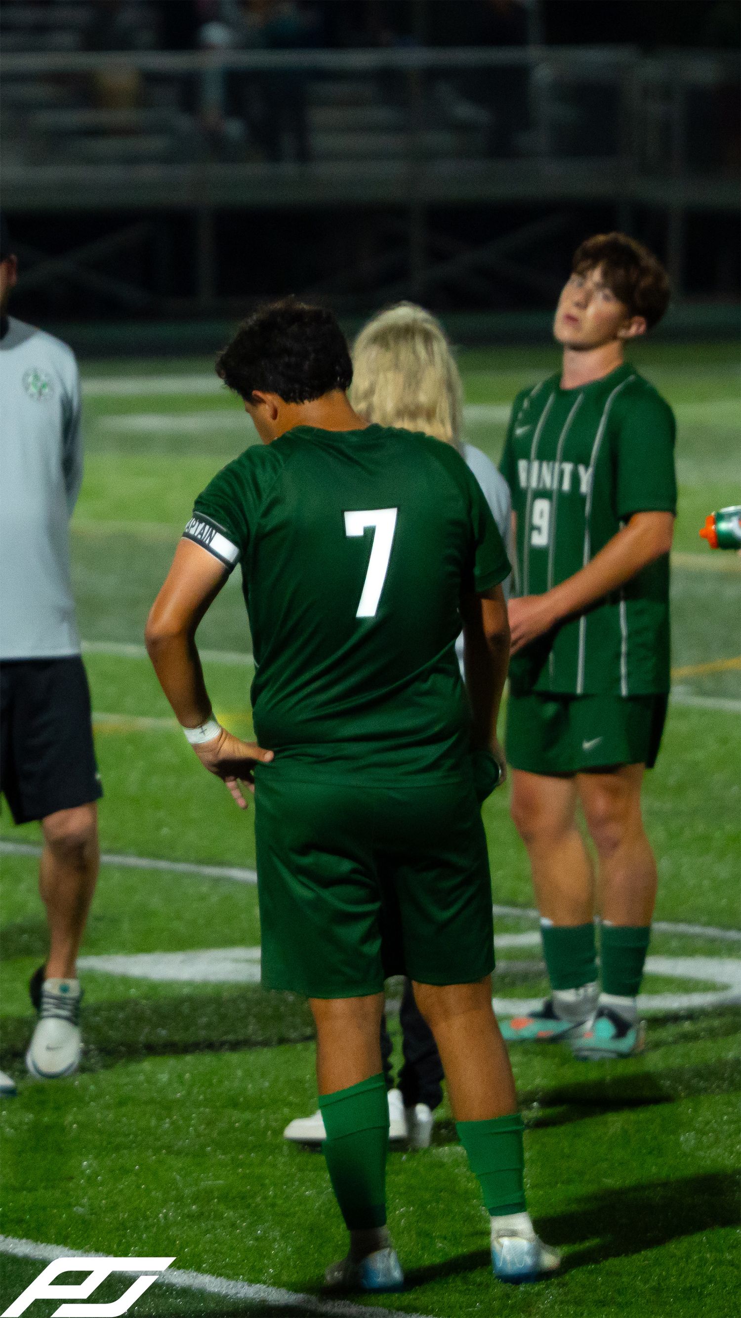 Two Trinity soccer players #7 and #9 in green uniforms stand on the sideline during a night game.