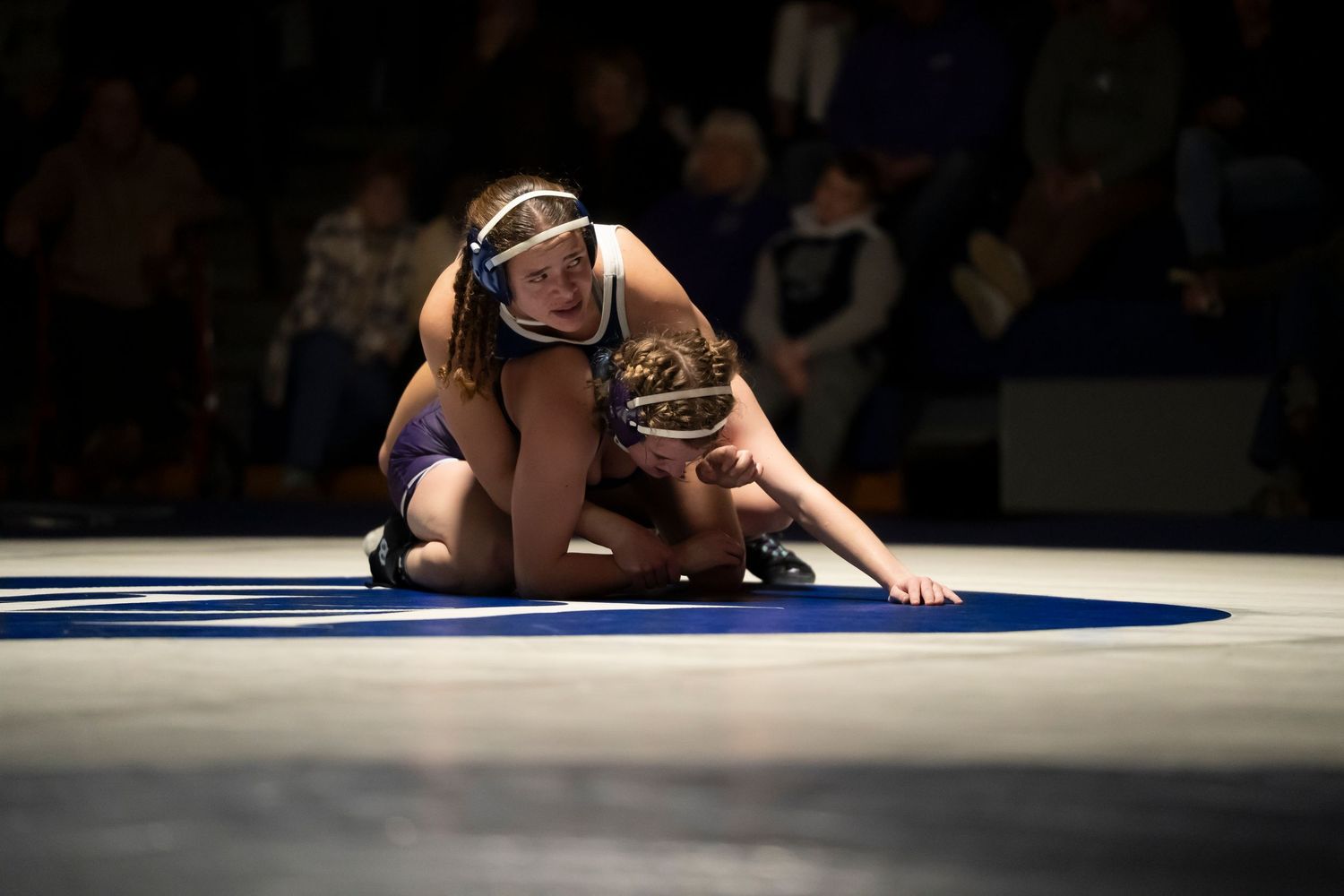 Female wrestler in blue singlet controls opponent on the mat during a high school wrestling match.