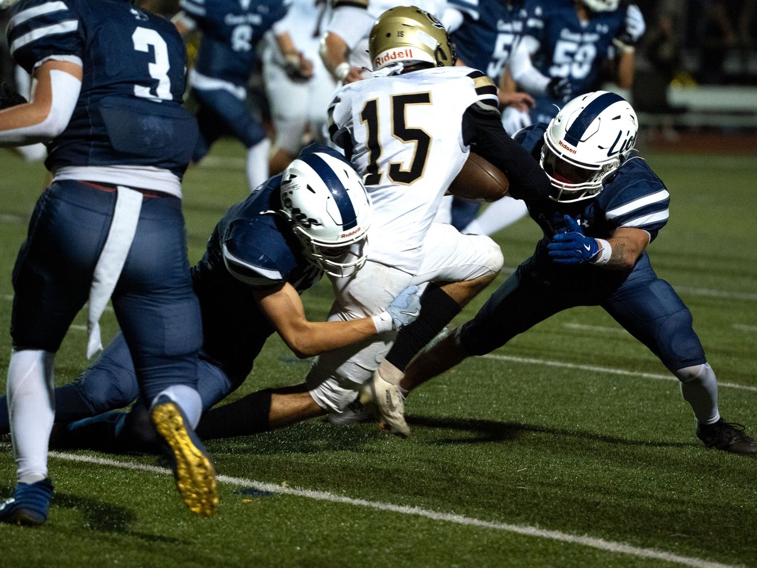 Two football players in navy uniforms tackle opponent wearing number 15 white jersey during night game.