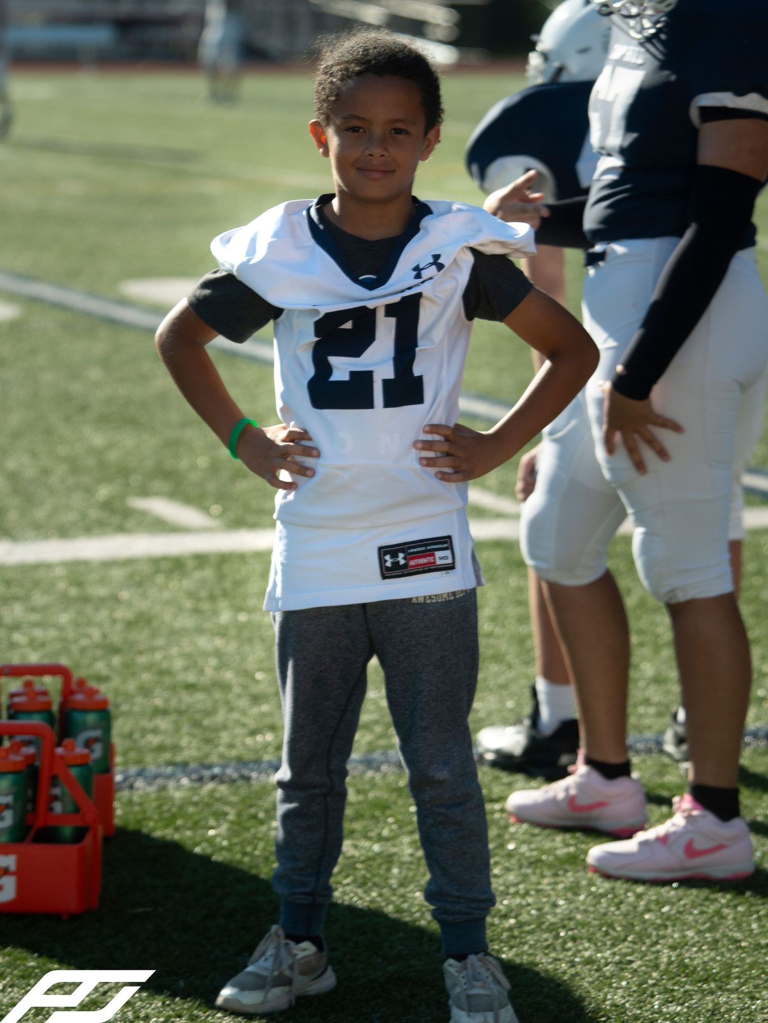 Child in Under Armour football jersey #21 stands confidently on sideline during youth football game.