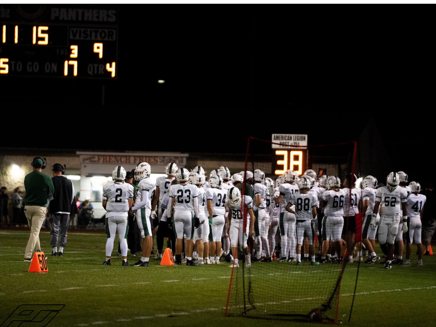 Football team huddles on sideline with lit scoreboard visible during night game.