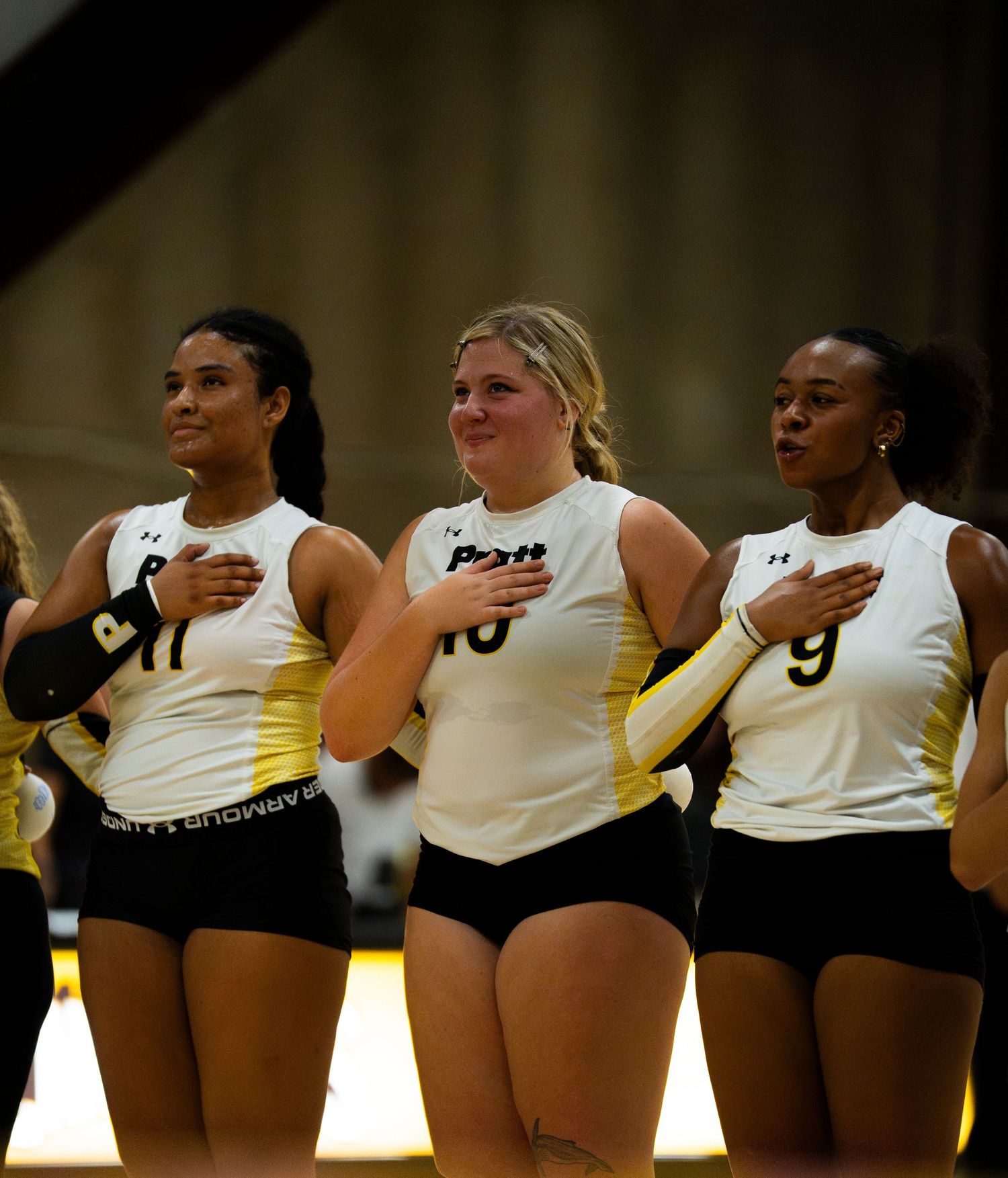 Three volleyball players in white and gold uniforms stand with hands over hearts during the national anthem.