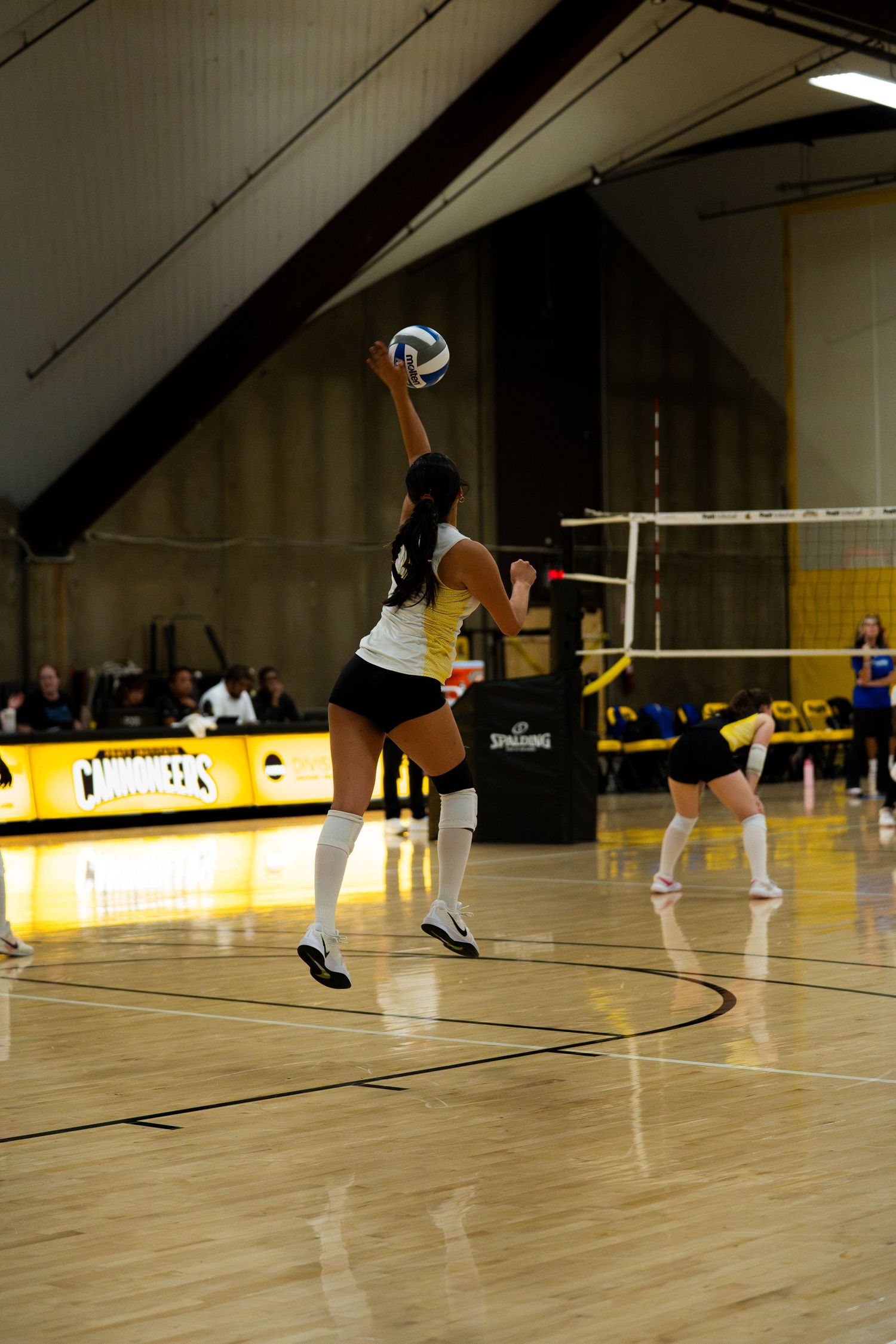 Female volleyball player jumping to serve indoors, teammate and net visible on polished hardwood court.