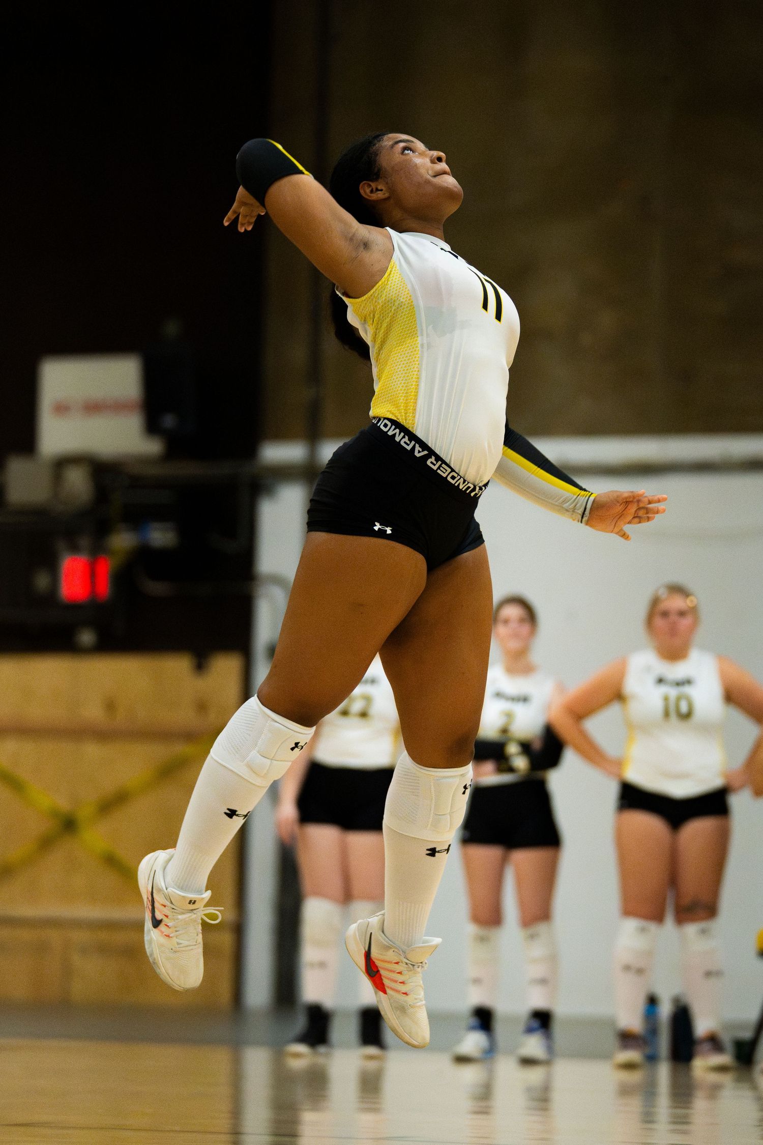 Female volleyball player in yellow and white uniform jumping to spike the ball during an indoor match.
