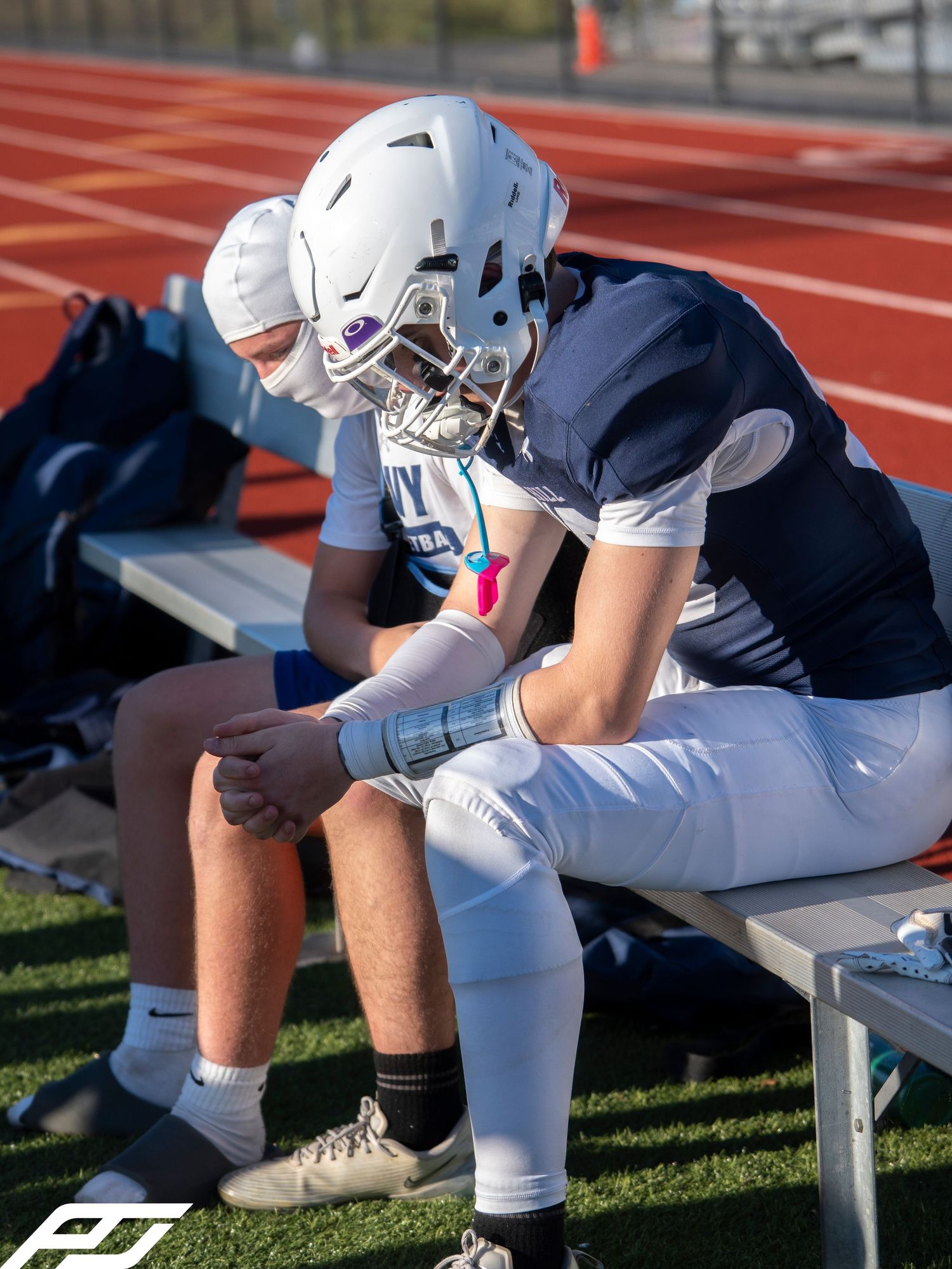 Two football players in navy uniforms sit on bench with helmets on during game.