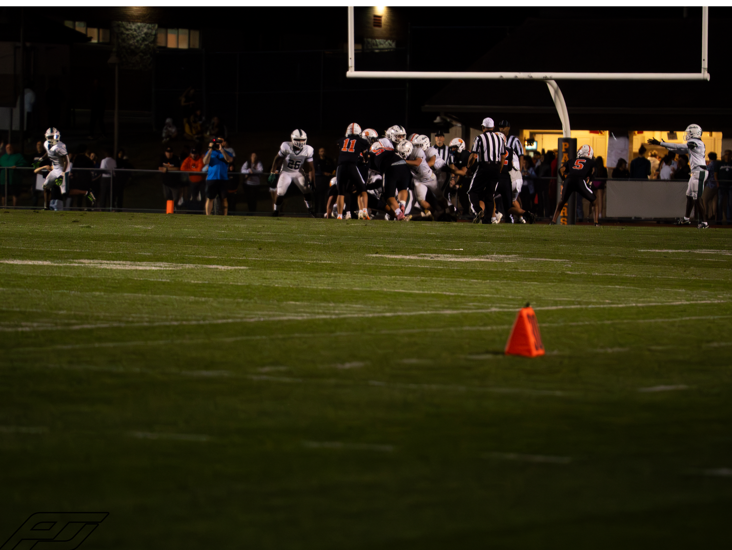 Night football game action with players in white uniforms competing on a green field with an orange cone marker.