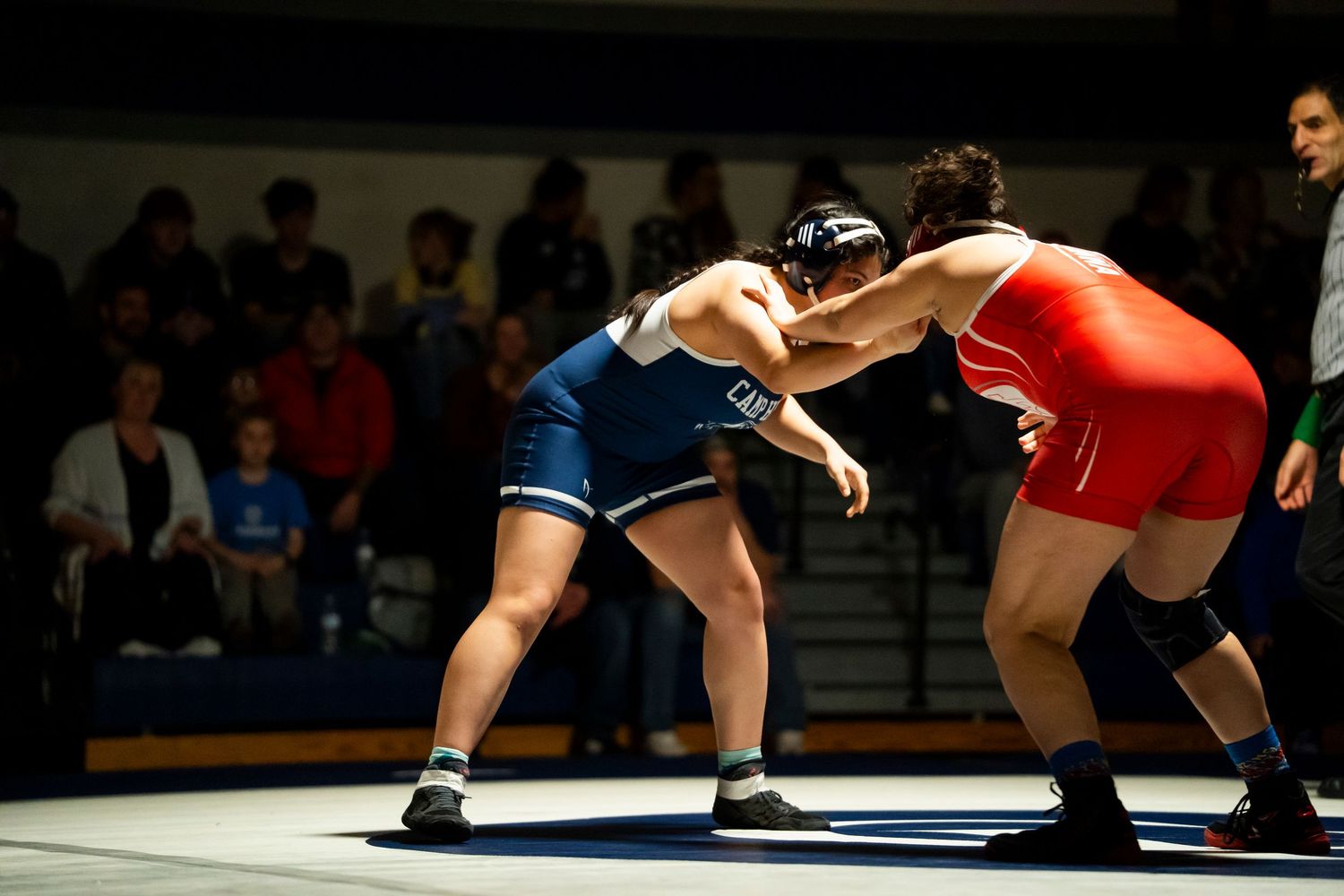 Two wrestlers in red and blue singlets engage in standing grapple during high school wrestling competition.