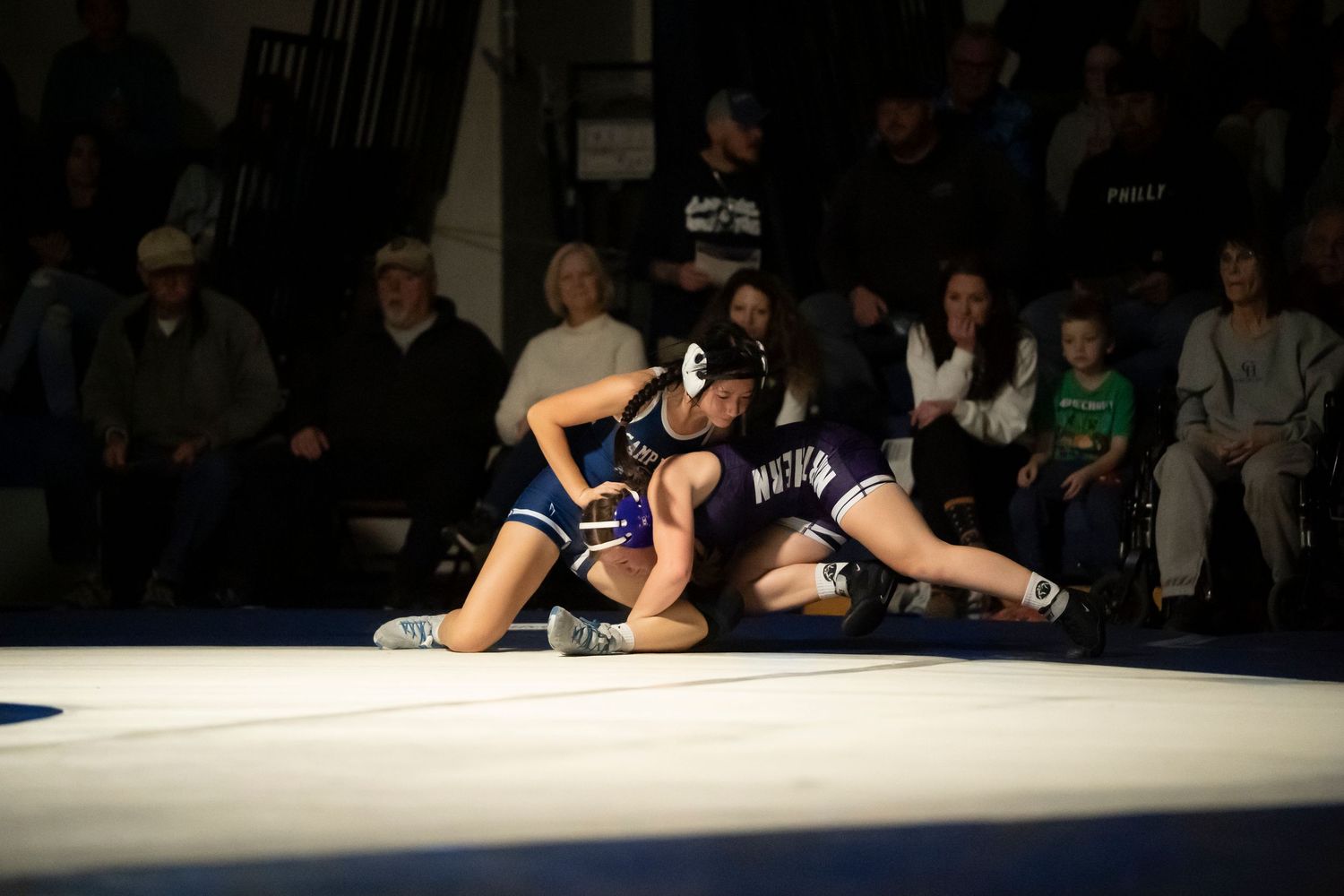 Two high school wrestlers compete on a blue mat with an audience watching in the background.