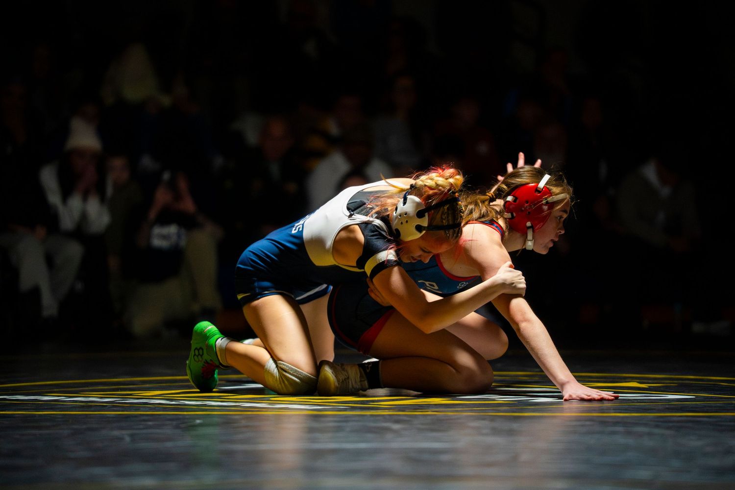 Two female wrestlers compete in intense grappling match on dimly lit wrestling mat.