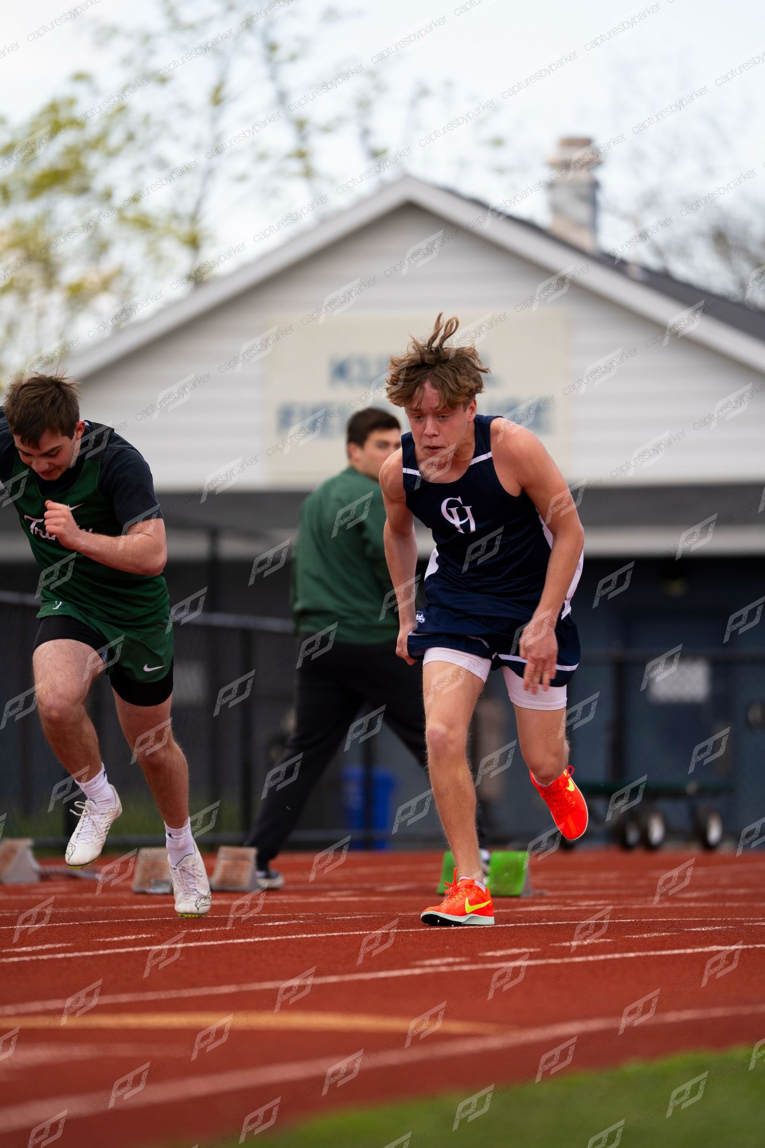 Male sprinter in GH blue uniform bursts from starting blocks on red track during outdoor meet.