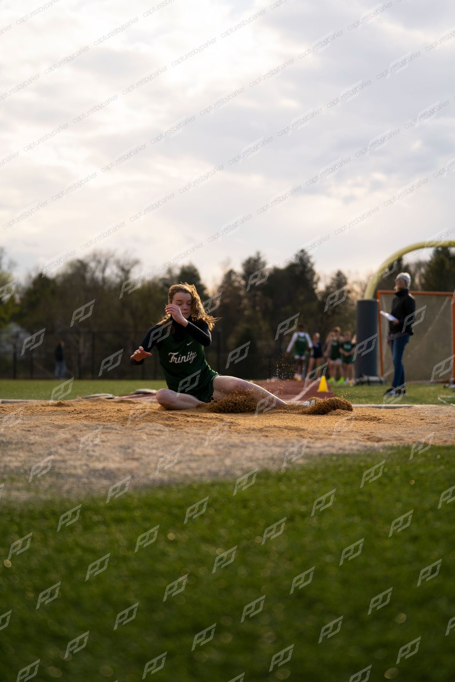 Female athlete in green Trinity uniform lands in sand pit during long jump competition.