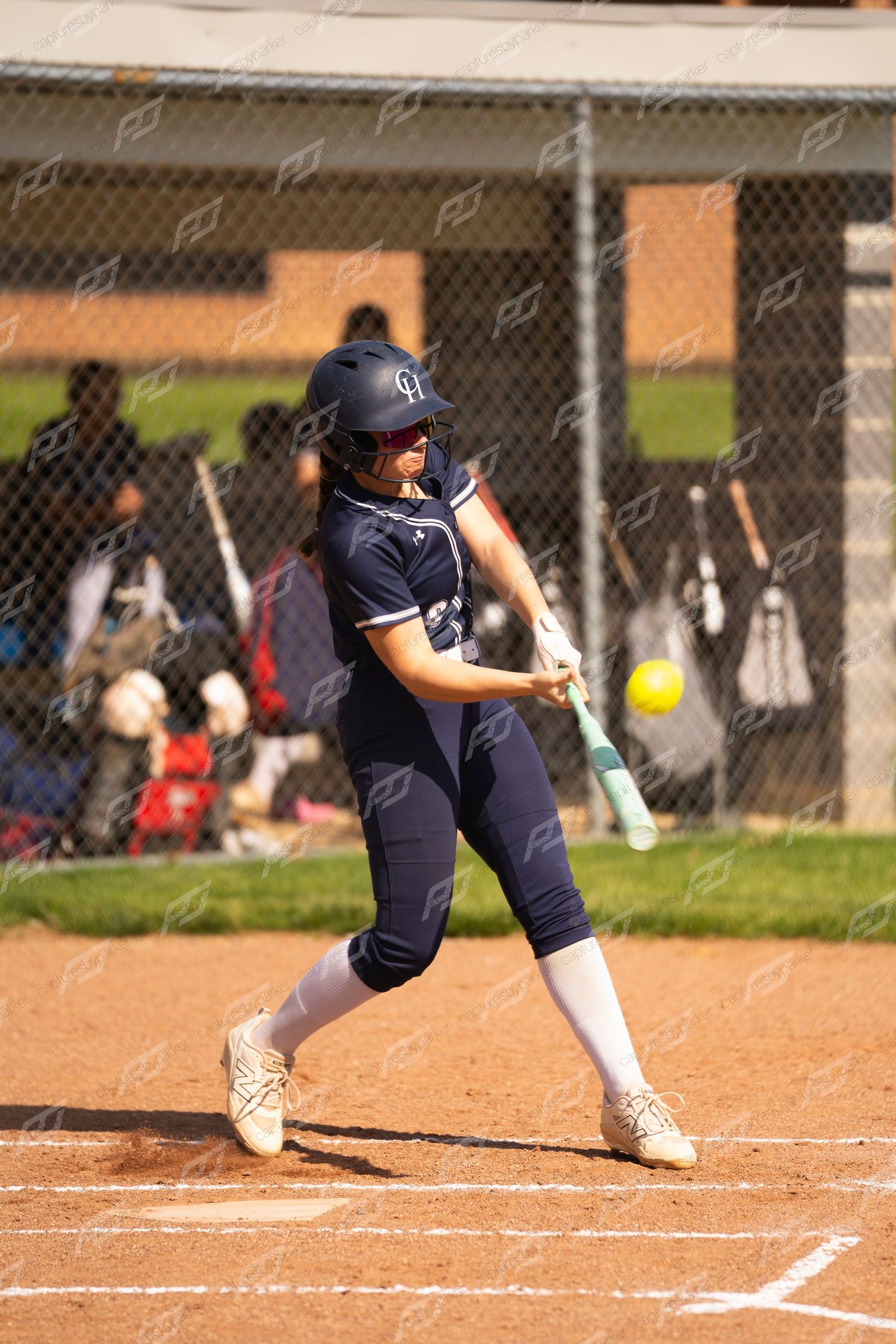 Female softball batter in navy uniform makes contact with yellow pitch using teal bat.