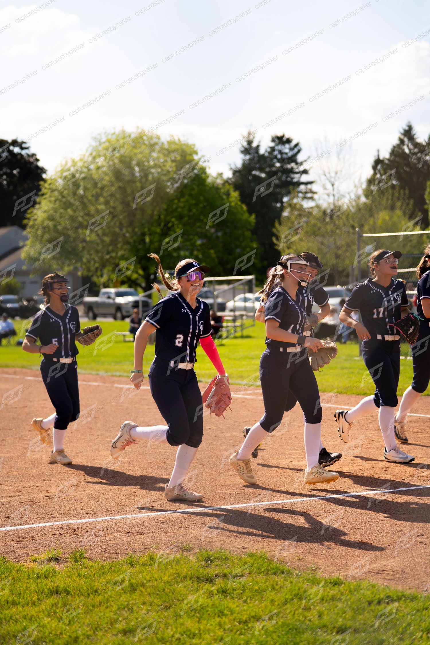 Smiling softball players in dark uniforms jogging across the field during a sunny game.