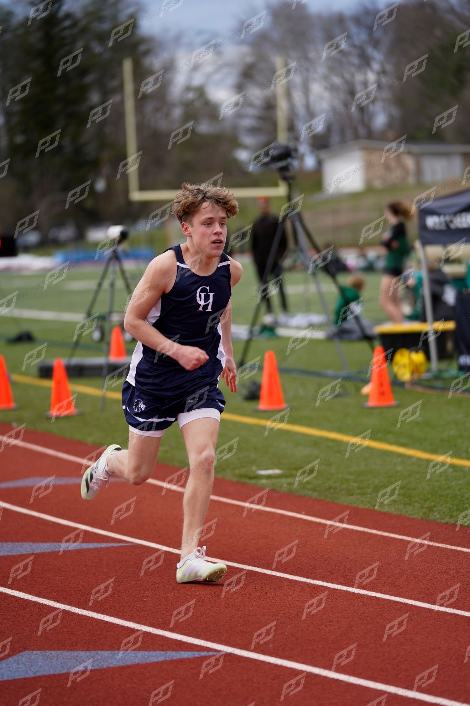 Male track athlete in navy CH uniform rounds a curve on red outdoor track during a competitive race.