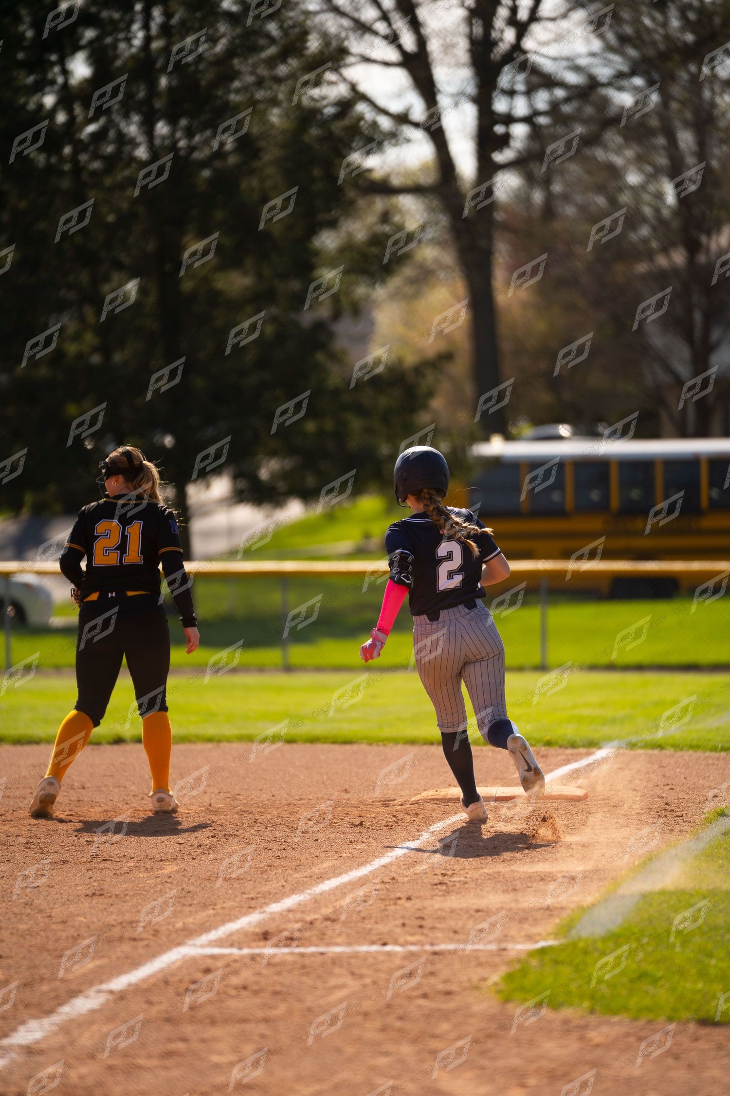 Softball player number 2 in navy uniform runs toward first base as opponent in yellow stands ready.