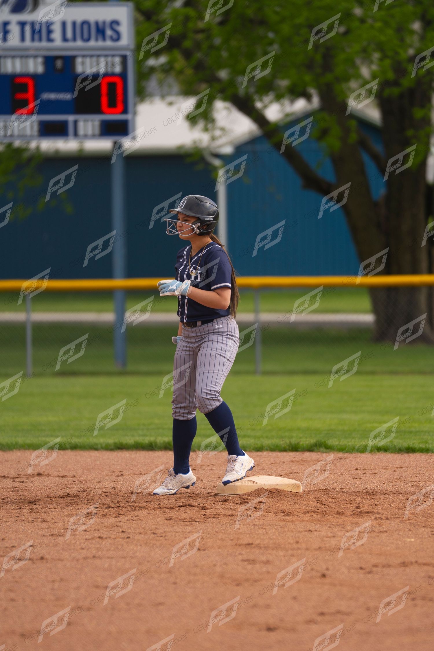 Softball player in navy pinstripe uniform stands on base with 3-0 scoreboard visible behind her.